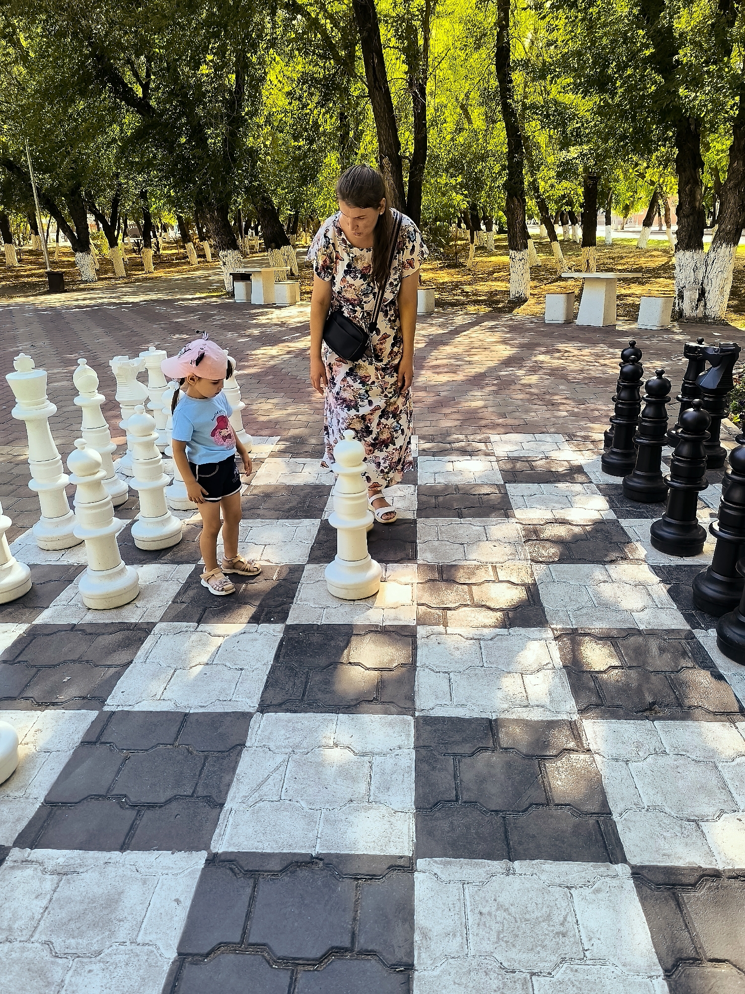 A woman and a young child stand on a giant chessboard in an outdoor park. The chessboard has large white and black pieces. The woman is wearing a floral dress and a black crossbody bag. The child is wearing a pink hat, a blue t-shirt, and dark shorts, and is standing to the left of a white pawn piece. In the background, there are trees with white-painted bases and benches. The scene appears to be set in Karaganda, Kazakhstan, as indicated by the city's typical park landscaping.