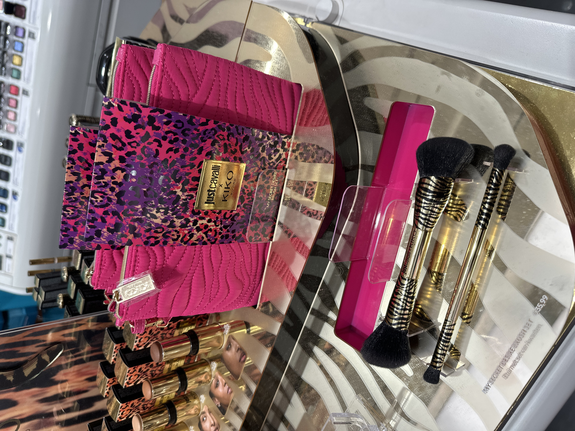 The image shows a display of Just Cavalli x Kiko Milano makeup products. The products include a pink quilted makeup bag, a leopard print eyeshadow palette, lipsticks, and a brush set labeled "My Secret Desire Brush Set." The brush set is priced at €35.99. The setting appears to be a retail display, possibly within a cosmetics store in Amsterdam, Netherlands.
