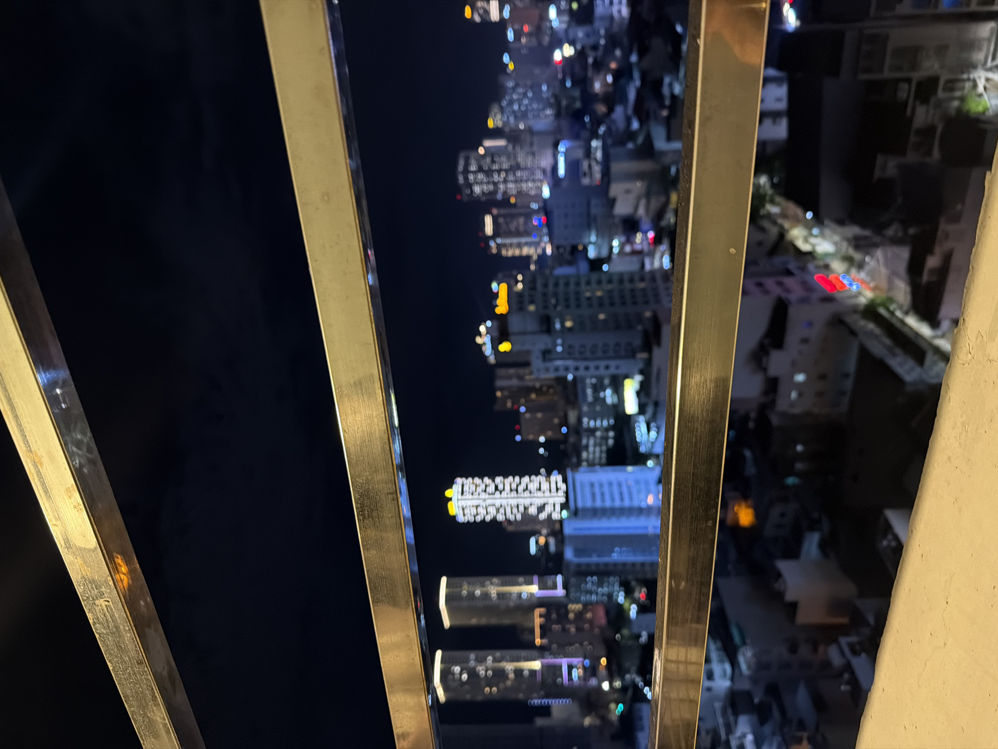 An eye-level shot looking down between balcony railings at night in Nha Trang Ward, Vietnam. Buildings with lit windows are visible through the bars and below the balcony in the distance. In the immediate foreground, a few dull metallic gold balcony railings frame the view. To the left of the image is a solid dark area. The cityscape has numerous high-rise buildings with illuminated windows. A mix of streetlights and vehicle lights, including red and white, dot the distant view. The railings are slightly out of focus, while the cityscape has a soft, dreamlike quality. The overall setting is an elevated perspective from a high-rise building at nighttime.