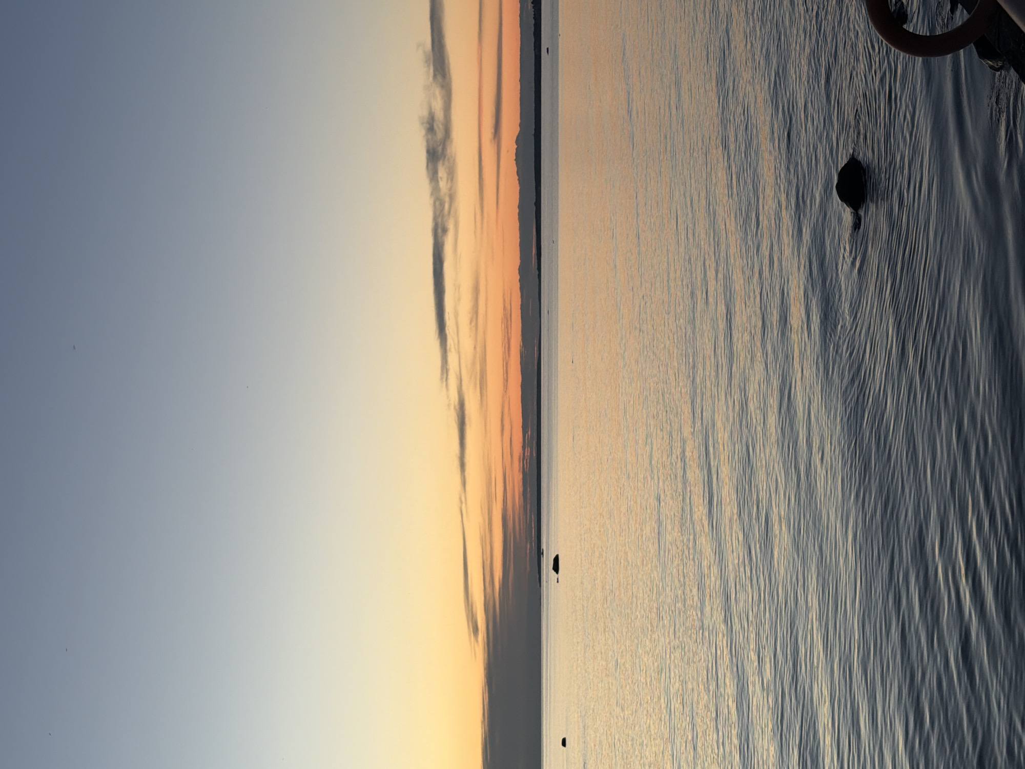 The image depicts a serene seascape at dusk, likely in Tallinn, Estonia, with the sky transitioning from a pale blue to hues of orange and yellow. The calm water reflects the soft colors of the sunset. Several dark rocks are visible emerging from the water at various distances. In the foreground, a portion of a reddish-orange structure, possibly part of a dock or railing, is visible on the right edge of the frame. The overall impression is one of tranquility and natural beauty during the twilight hours.