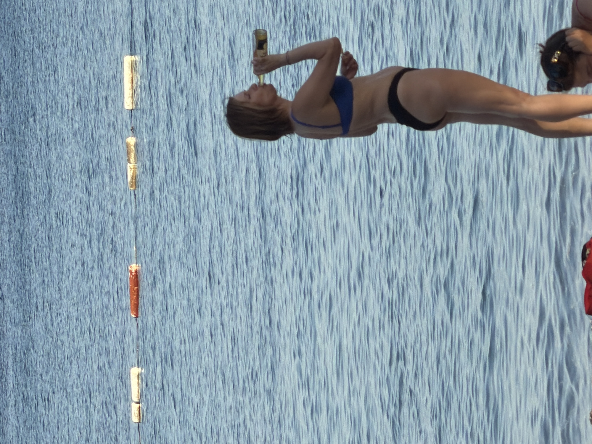 A daytime outdoor shot depicts a woman in a blue bikini top and black bottoms drinking from a bottle, with another woman partially visible to the right, on what appears to be water, possibly the sea. To the left, vertical markers are visible, attached to a vertical cable. The setting may be coastal, potentially related to Budva, Montenegro. The blue-toned texture in the background suggests the water surface.