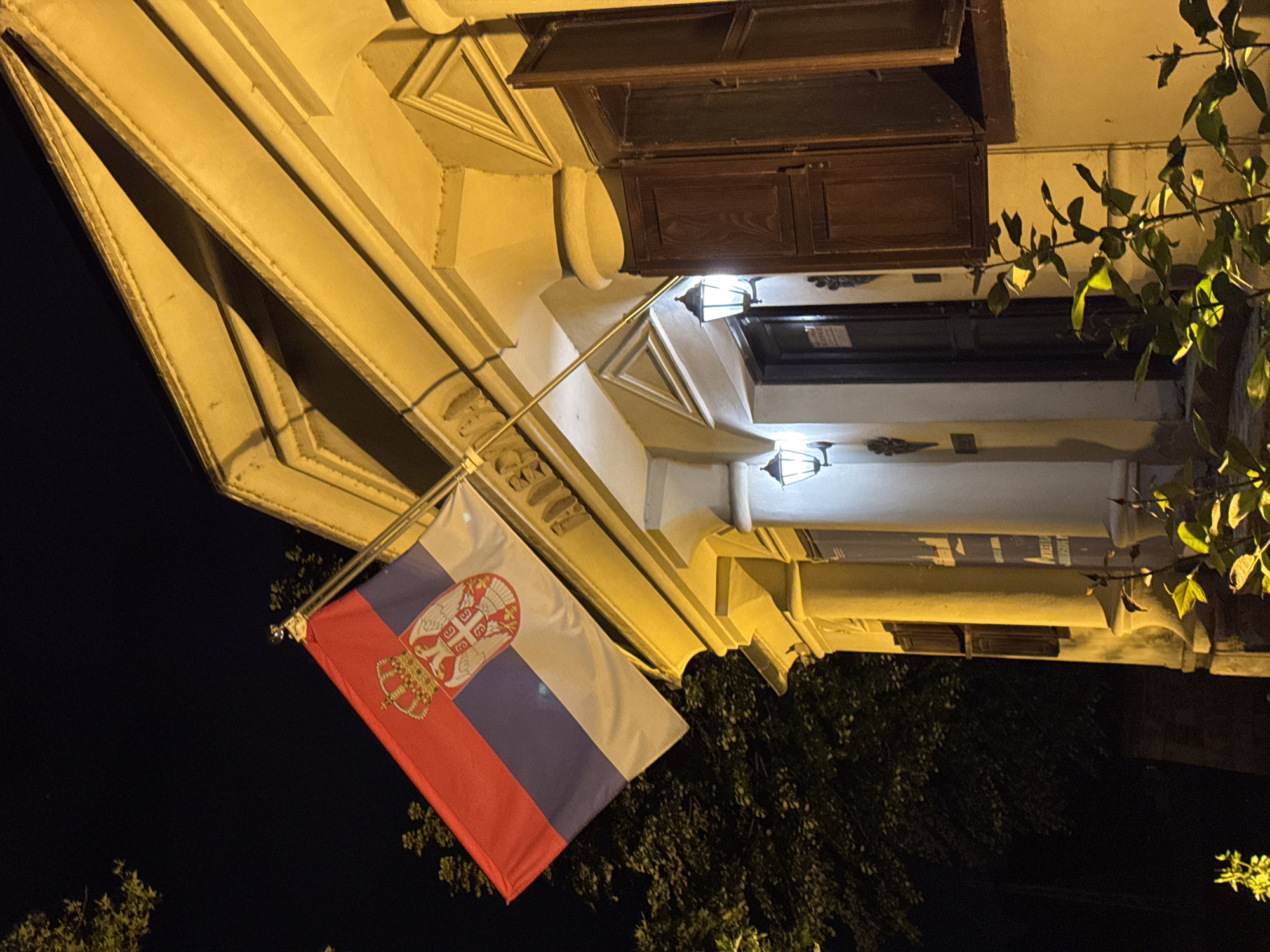 The image shows a building with the flag of Serbia hanging from it. The building has a classic architectural style with detailed moldings and a window with dark wood panels. Two ornate light fixtures are visible above a doorway. The building appears to be illuminated by artificial light, casting a yellow hue. Part of a tree with green leaves is visible on the right side of the building. The background is dark, suggesting it is nighttime. Based on the architectural style and the presence of the Serbian flag, it is likely the image was taken in Belgrade, Serbia. The exact location in the city is not discernible from the image alone.