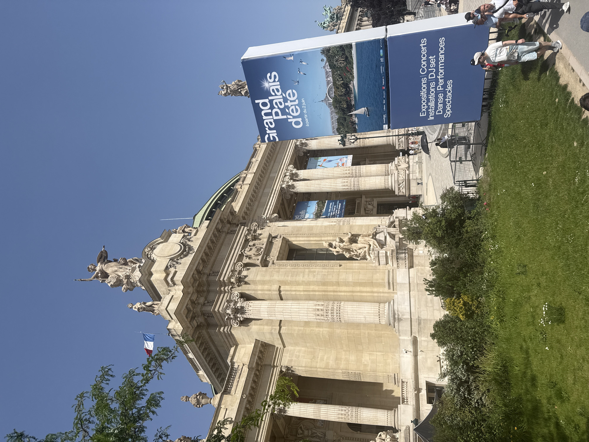 The image shows the Grand Palais d'été in Paris, France. It is a large, ornate building with columns and statues. There is a large sign in front of the building advertising the summer events, which include exhibitions, concerts, installations, DJ sets, dance performances, and shows. In the background, people are standing and sitting nearby, and a French flag is visible at the top of the building. There are trees and grass in the foreground.