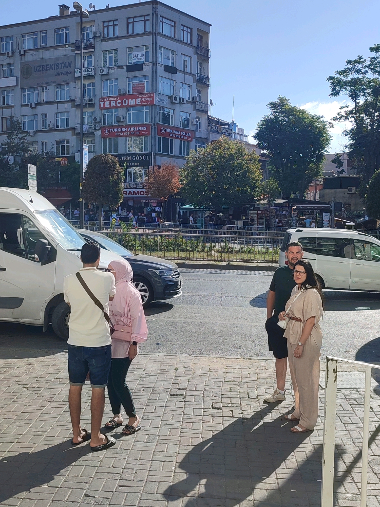 The image captures a street scene in Istanbul, Turkey, on a sunny day. In the foreground, four individuals stand on a paved sidewalk. To the left, a man wearing a white t-shirt, dark shorts, and dark slides with a cross-body bag, faces away from the camera. Next to him, a woman in a pink hijab and a light pink outfit, with a pink purse, faces towards the man. To the right of the couple, a man in a dark green t-shirt and black shorts, and a woman in a beige jumpsuit and white sandals, face the camera. They are positioned near a white van and a dark sedan, with a metal railing in the far right foreground.
Behind the people, a multi-story building with numerous windows and air conditioning units is visible. Advertisements, including "UZBEKISTAN airways" and "TURKISH AIRLINES," are displayed on the building's facade. Further back, trees and other buildings form the urban landscape, with a clear blue sky overhead. The scene suggests a public urban environment with moderate pedestrian and vehicular activity.