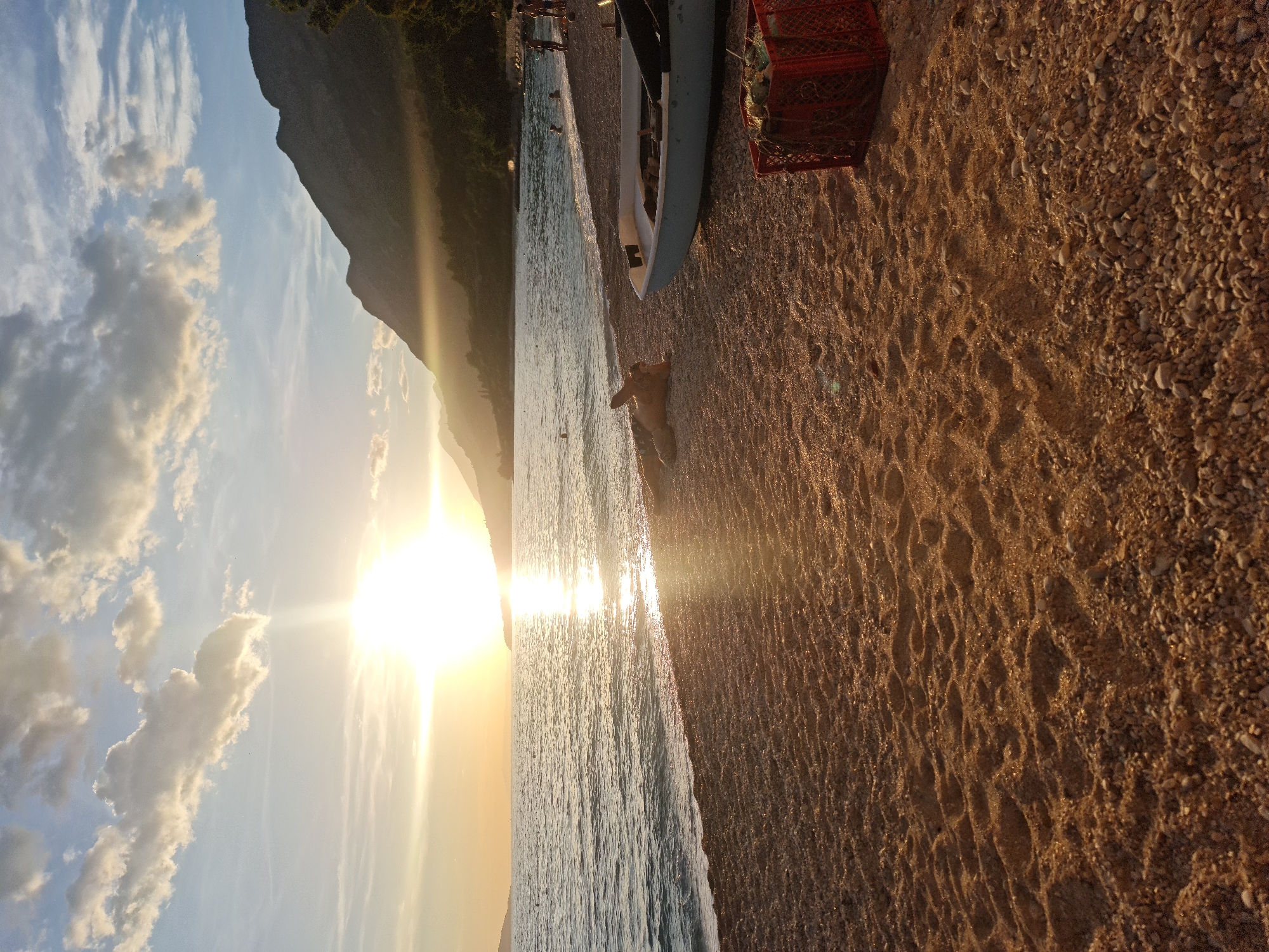 The image depicts a tranquil beach scene during sunset in Općina Gradac, Croatia. The sun, a bright orb with visible rays, is low on the horizon, casting a warm glow across the sky and water. The sky is adorned with scattered clouds, some tinged with orange and pink hues from the setting sun.
The foreground is dominated by a pebble and sand beach, with distinct footprints and ripples in the sand. To the right, a red plastic crate is placed on the beach, containing some green foliage. Beside it, a light-colored boat is partially visible, resting on the shore. In the middle ground, a person is lying down on the beach, appearing relaxed.
The sea stretches out towards the horizon, with gentle waves lapping at the shore. The water reflects the golden light of the sunset, creating a shimmering path. In the distance, a hilly coastline is silhouetted against the bright sky, with some trees visible along its slopes. A few other people are scattered along the water's edge and in the sea, enjoying the evening. The overall atmosphere is peaceful and serene, characteristic of a Mediterranean sunset.