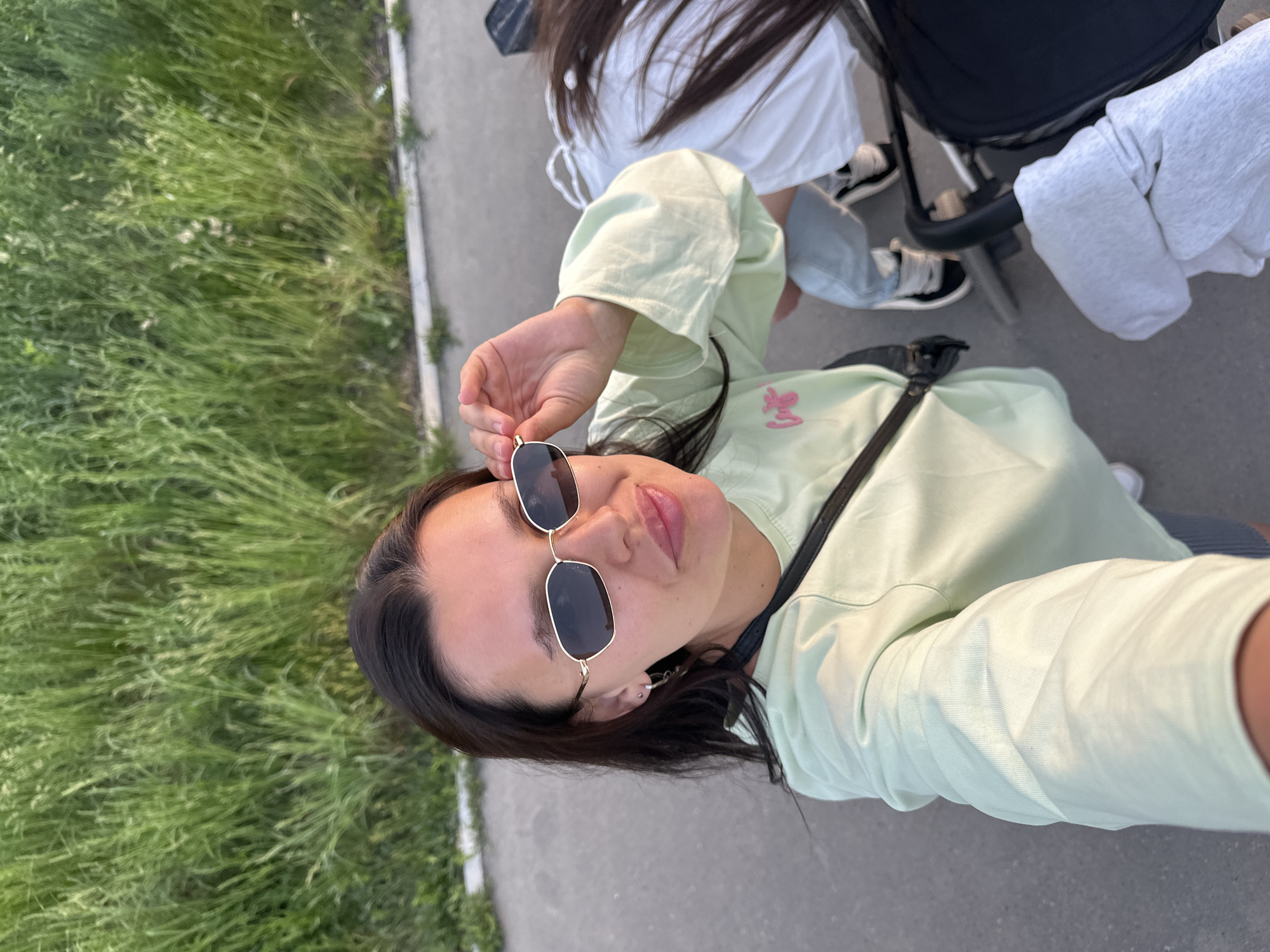 A selfie photograph shows a woman wearing a light green shirt and black sunglasses standing on a paved surface with a patch of green grass visible to the left. The woman is holding up her sunglasses with her right hand. To her right, partially visible, are a person in a white shirt, light-wash jeans, and black shoes, and a black baby stroller covered with a white fabric. The location is Chaikovsky, Russia.