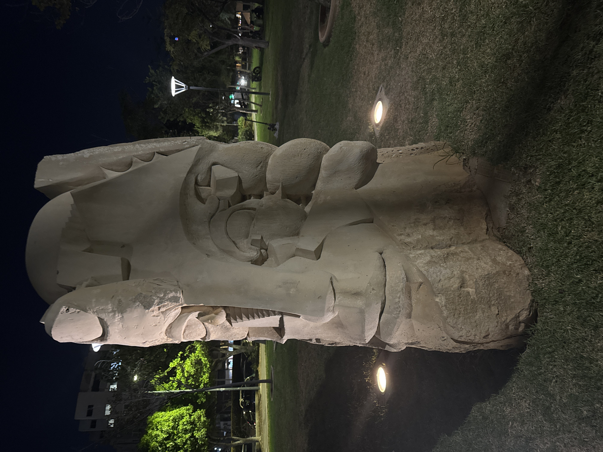 The image captures a large, abstract stone sculpture in an outdoor park at night. The sculpture is roughly humanoid in shape, resembling a face with stylized features like a nose and mouth, carved from a rough, light-colored stone. It stands tall on a patch of green grass, illuminated by embedded ground lights that cast warm pools of light and long shadows. In the background, trees with vibrant green foliage are visible, suggesting a well-maintained park. Beyond the trees, there are buildings with lights on, indicating an urban setting. A lamppost with a modern design stands to the right of the sculpture, also emitting light. The overall impression is of a public space at night, with artificial lighting highlighting the art installation and the surrounding greenery. The specific location is not identifiable from the visual cues in the image.