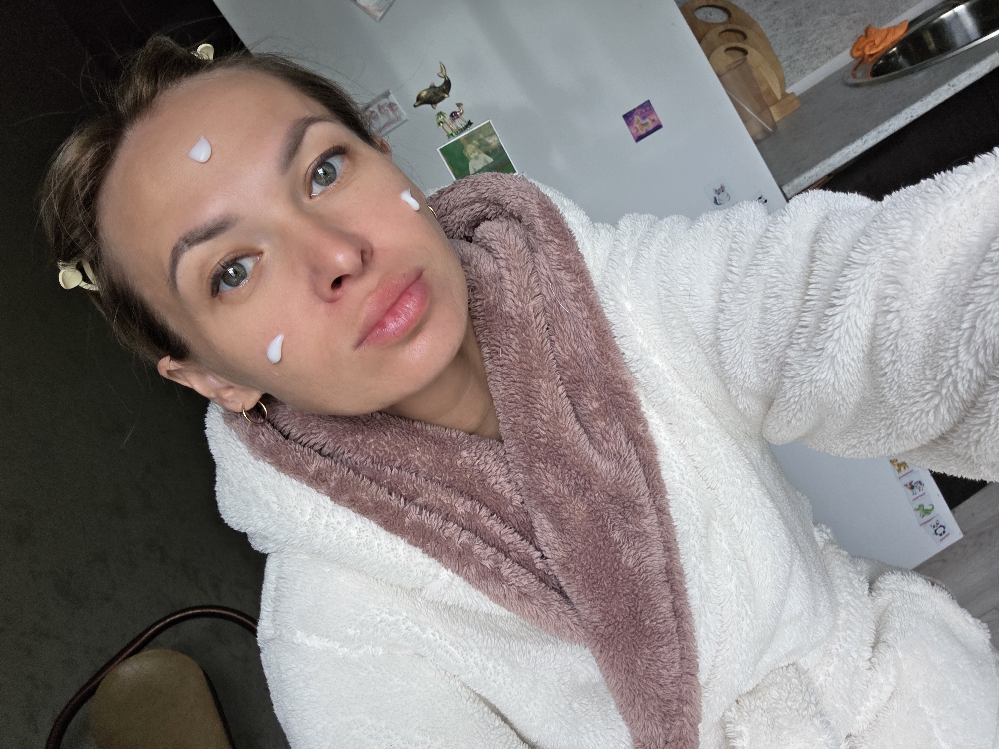 A woman in a cozy, fluffy bathrobe takes a selfie in what appears to be a home in Chaikovsky, Russia. She is facing the camera, holding it slightly above eye level, capturing her upper body and a glimpse of her surroundings. Her light brown hair is pulled back, secured by several small, light-colored claw clips. She has noticeable drops of white or off-white cream or lotion on her forehead, left cheek, and nose, suggesting she is in the process of applying skincare. Her eyes are a striking light green or hazel, and her lips are naturally full with a slight sheen. She wears a simple gold hoop earring in her visible ear.
The bathrobe is predominantly white and very textured, with a soft, plush material. The inner lining, visible around the collar and lapel, is a muted pink or dusty rose color. The background reveals an indoor setting, likely a kitchen or an area adjacent to one. To the right, a white refrigerator is visible, adorned with small, colorful magnets or stickers. Above the refrigerator, a wooden holder for kitchen utensils and a dark grey or black countertop with a metal basin (possibly a sink) and an orange cloth are present. More decorative elements, including a golden dolphin figure, a small picture frame, and other small wall decorations, are on the wall behind her. The overall impression is one of a relaxed, domestic moment focused on self-care. No clear, legible text is visible on any objects.