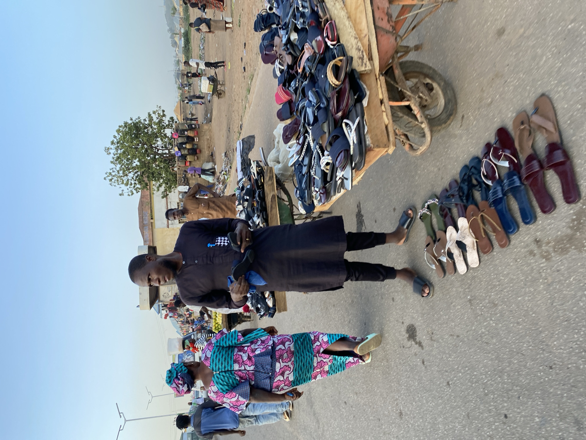 The image shows a street scene in Bukuru, Nigeria. A man in a black shirt and pants is holding a pair of sandals, apparently selling them. A woman wearing a colorful, patterned dress and head wrap is standing in front of him, likely a customer. A cart filled with sandals sits beside them, and more sandals are laid out on the ground for display. Other people are visible in the background, along with buildings and a tree. The scene takes place on a paved road with some trash on the side, indicating a street market or informal selling area.