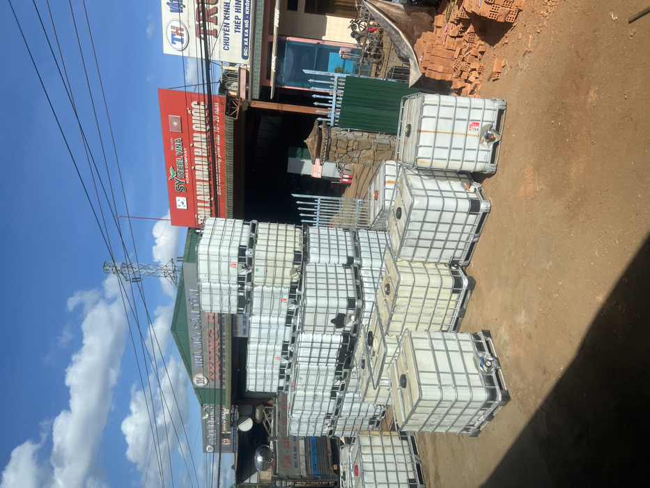 The image shows a stack of intermediate bulk containers (IBCs) in front of two businesses. One sign reads "SY STEEL VINA KOREA COMPANY" and another is "NHÀ MÁY CÁN TÔN-SA." The containers are caged in metal frames. Overhead power lines are visible against a blue sky with clouds. Bricks and other materials are visible beyond the IBCs. The environment appears to be an industrial or commercial area.