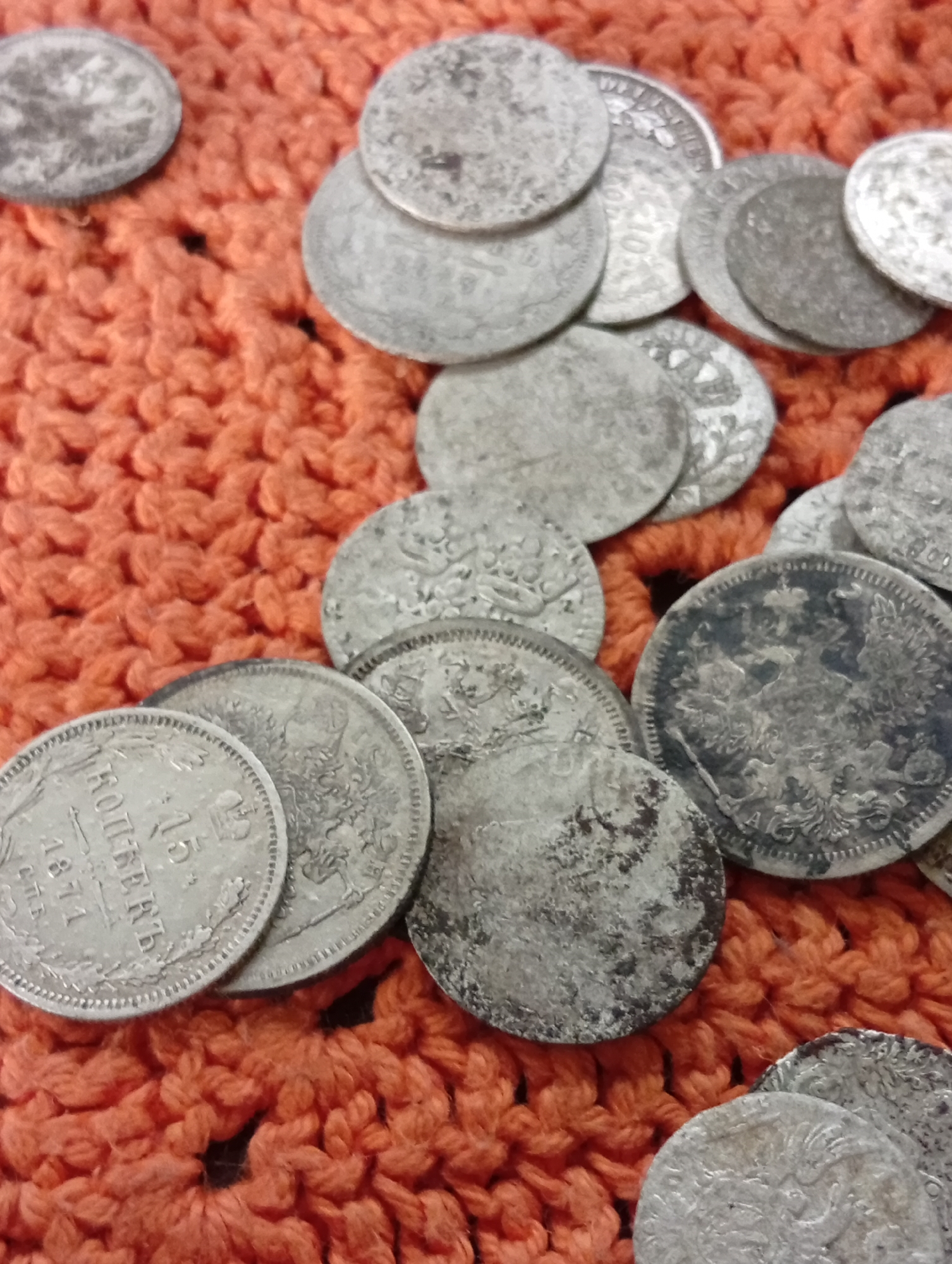 A pile of antique silver coins, varying in size and condition, are scattered across an orange crocheted surface. The coins exhibit intricate designs and some show signs of wear and tarnish. One coin in the foreground, dated 1871, displays Cyrillic lettering. The crocheted material has a textured, woven pattern. The lighting suggests an indoor setting, possibly a close-up studio shot. The location of Varputėnai, Lithuania, is not discernible from the visual information provided in the image.