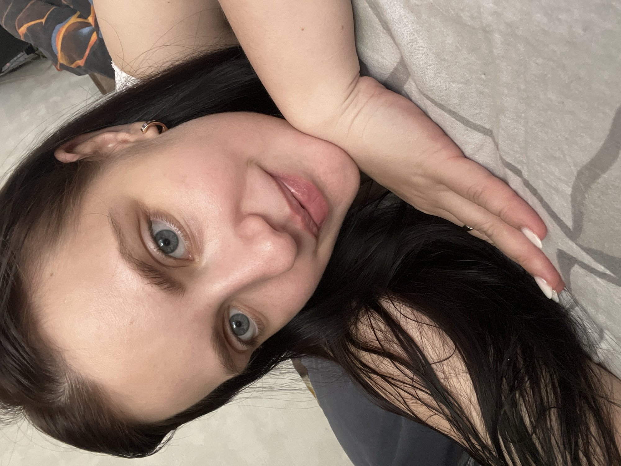 A selfie image shows a fair-skinned person with dark hair and light blue eyes, lying down. The person's head is turned slightly towards the viewer, with their hand resting against their face. The background includes a light gray, textured surface, possibly bedding. The specific location within Марковский, Russia, is not discernible from the image.
