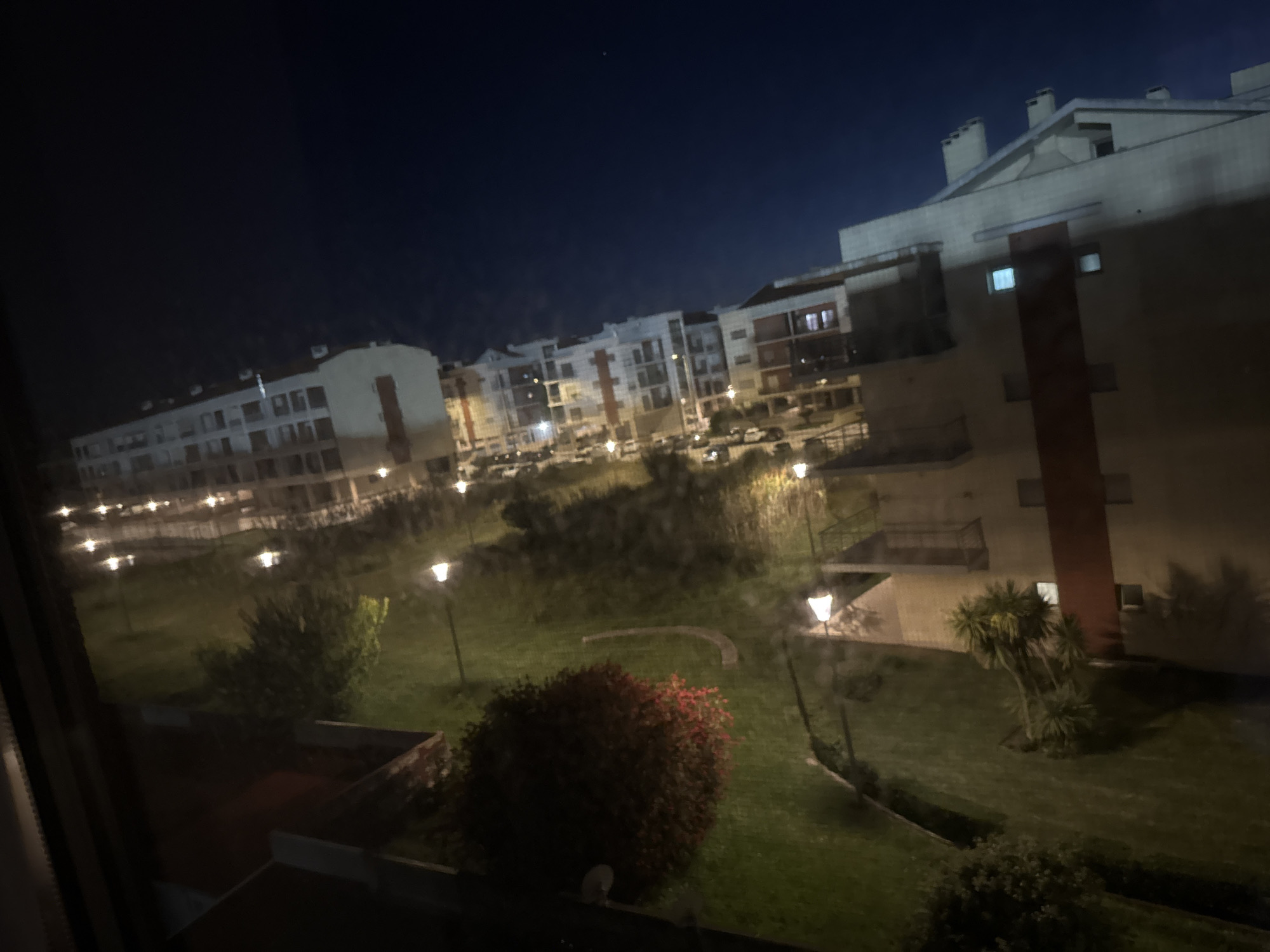 The image depicts a night scene in Figueira da Foz, Portugal, viewed from an elevated perspective, likely a window. Foreground shows a manicured garden with shrubs and grass. Multiple lit lampposts are scattered throughout the garden. Beyond the garden are residential buildings, some with visible windows. Cars are parked on the streets between the buildings. The night sky is dark, with a single visible star. The location appears to be a residential area, possibly near the coast, based on the city's geographic location. No people are visible.