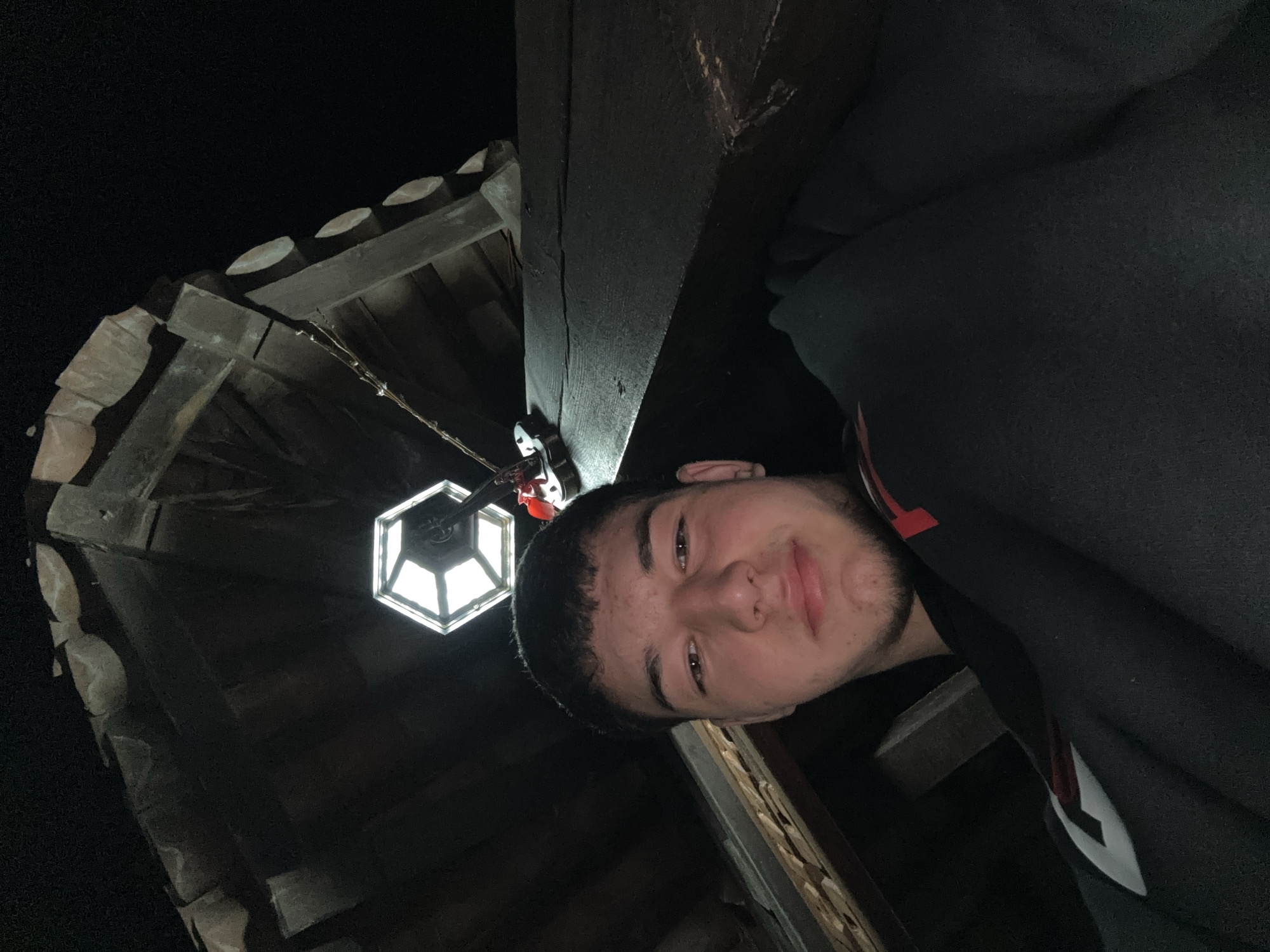 A young man poses for a photo, looking directly at the camera with a neutral to slight smile. He has dark hair, light facial stubble, and is wearing a dark hooded garment with visible red and white detailing on the shoulder or arm. The image is taken from a low angle, rotated slightly.
The scene is dimly lit, indicating it is nighttime. A hexagonal lantern with illuminated panes hangs above and to the left of the man, casting light on his face and the surrounding structures. Above and behind the man, a wooden roof structure is visible, supported by dark wooden beams. The roof features traditional, curved tiles, likely made of terracotta or stone, which are partially obscured by shadow but detailed in the accompanying crops. The background is dark and indistinct, emphasizing the illuminated foreground.
The setting appears to be an outdoor or semi-outdoor space, possibly under a covered porch or within a traditional architectural structure, consistent with the location context of Zlatograd, Bulgaria, which is known for its preserved old town and traditional buildings. There is no visible text in the image.