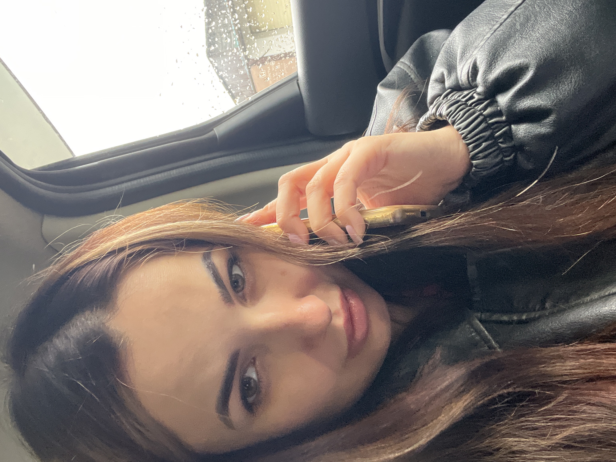 An adult female is captured in a selfie taken from inside a vehicle, occupying the left and central portion of the frame. She has long, dark brown hair with lighter, possibly auburn, highlights, parted down the middle and falling over her shoulders. She wears a black leather jacket with an elasticized, ruched cuff visible on her right arm. Her right hand, with light pink nail polish, holds a gold-toned smartphone to her right ear. She looks directly into the camera with a focused expression. Through the car's right passenger window, numerous raindrops are visible on the glass, suggesting wet weather conditions. Beyond the window, the outdoor environment is blurred, showing an overcast sky and the indistinct outlines of a building structure featuring a light-colored wall and a dark roofline. Dark-colored car interior trim surrounds the window frame.