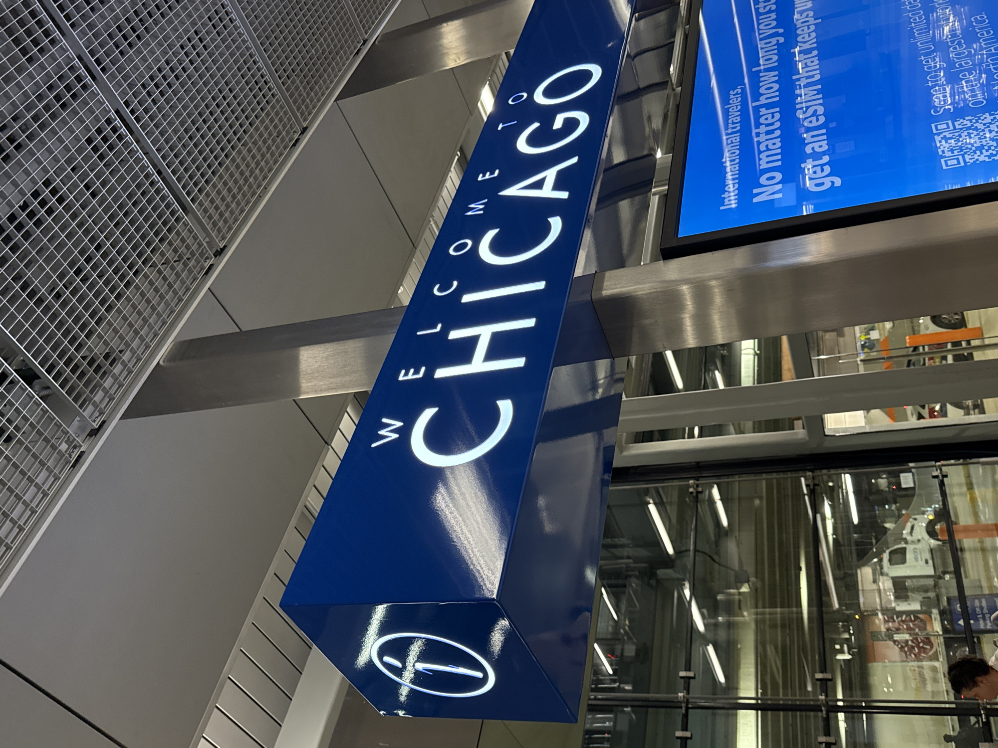 A low-angle, tilted shot presents a blue sign with "WELCOME TO CHICAGO" in white capital letters, hanging from the ceiling, likely at O'Hare International Airport (ORD) in Chicago, United States of America. A smaller circular logo is at the sign's bottom. Adjacent to the sign is a digital screen displaying travel information, a QR code, and the text "International travelers, No matter how long you stay, get an eSIM that keeps..." Behind the sign and screen is a glass-enclosed area that reflects bright linear light sources and contains infrastructure, possibly an airport terminal or transport facility. At the bottom right of the frame, the back of a person's head is visible. Above the sign, there is a ceiling with a dense, square-patterned metal grid.