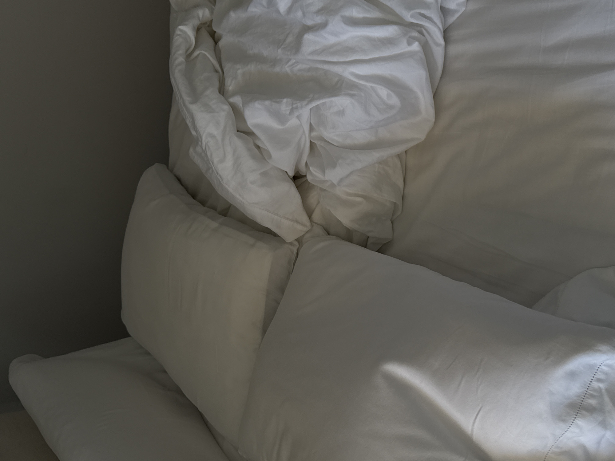 The image displays an unmade bed, viewed from a high angle, likely in a bedroom. The primary subjects are white bed linens, including several pillows, a fitted sheet, and a crumpled duvet or blanket. The bedding appears soft and has various folds and wrinkles, indicating recent use or an undisturbed state.
On the left side of the frame, a dark grey wall serves as a backdrop, providing a neutral contrast to the white textiles. No people are visible in the scene, and no specific activity is explicitly depicted. However, the disheveled appearance of the bedding suggests either recent occupancy or a preparation for rest.
Notable details include the lighting, which casts soft shadows and highlights across the white fabrics, particularly on the rightmost pillow where a strong highlight suggests a light source from the upper right, possibly an unseen window. This lighting implies either early morning or late afternoon, contributing to a calm and serene atmosphere. There are no visible location cues within the image itself that specifically point to Mexico City, Mexico, other than the provided context. No visible text is present in the image. The overall impression is one of comfort, relaxation, and tranquility within a simple, understated interior space.