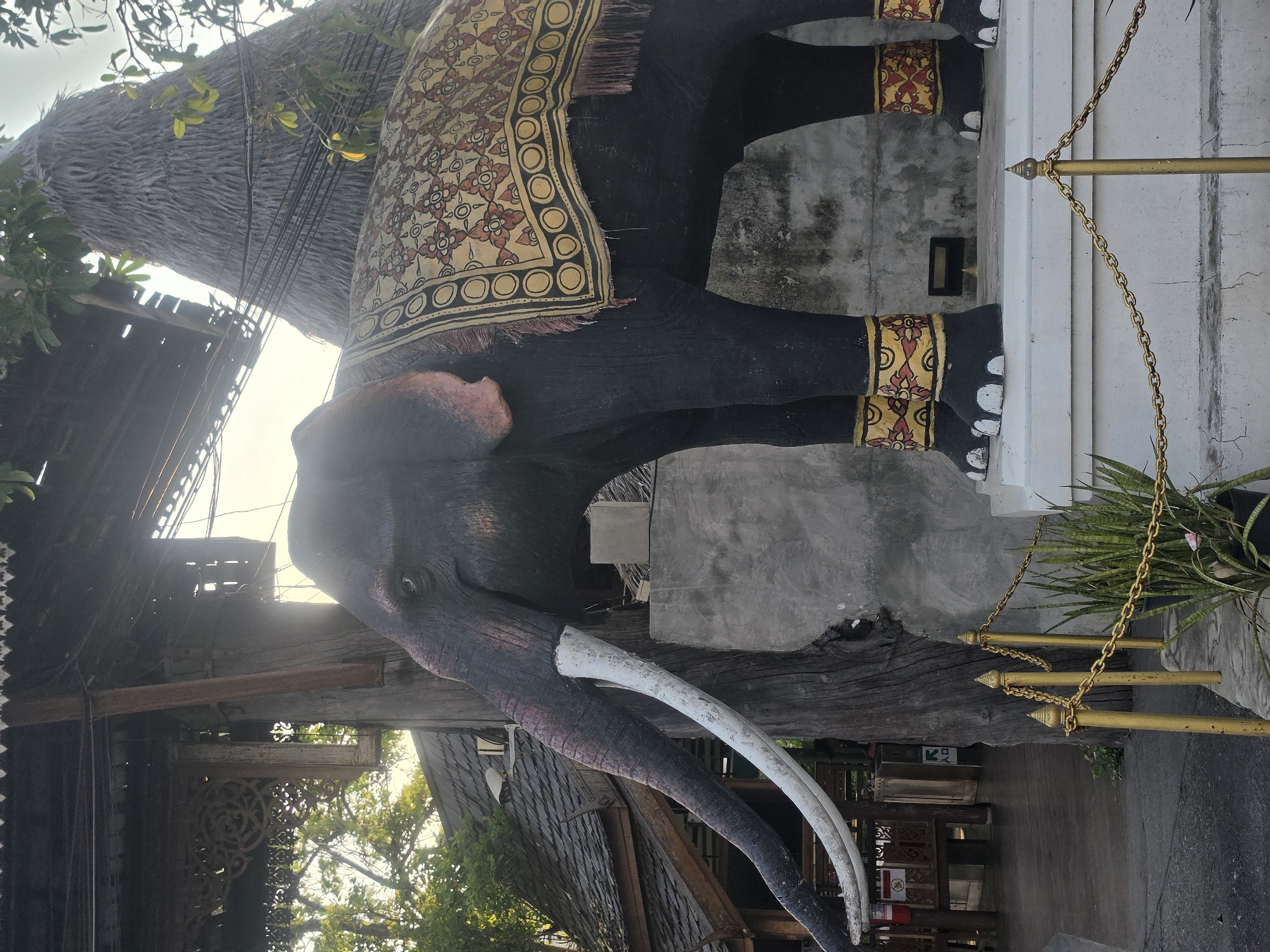 This image captures a large, intricately detailed elephant statue set in an outdoor environment, likely in Pattaya City, Thailand. The statue, oriented as if walking left, is predominantly dark grey or black with significant white tusks and a textured trunk. It is adorned with a decorative saddle blanket featuring a vibrant pattern of yellow, red, and black floral and geometric motifs. Similar ornamental bands wrap around the elephant's lower legs, which terminate in white-painted feet.
The elephant statue is positioned alongside a white, concrete-like structure, possibly a pillar or platform. A golden chain barrier, supported by golden posts with pointed finials, runs parallel to this structure, partially obscuring some green potted plants, possibly snake plants, behind it.
The background reveals architectural elements suggesting traditional Thai design, including a multi-layered, thatched or textured roof and dark wooden beams with carved details. Overhead, green foliage from trees is visible, contrasting with a bright, clear sky, indicating a daytime setting with fair weather.
Several textual elements are present. In the lower-left, a directional sign features a white arrow pointing left with the letters "KFC" underneath, suggesting proximity to a restaurant. In the lower-right corner, a "no smoking" sign, depicted by a red circle with a diagonal line over a cigarette, is partially visible. The overall scene conveys a sense of cultural aesthetics and a bright, serene atmosphere.