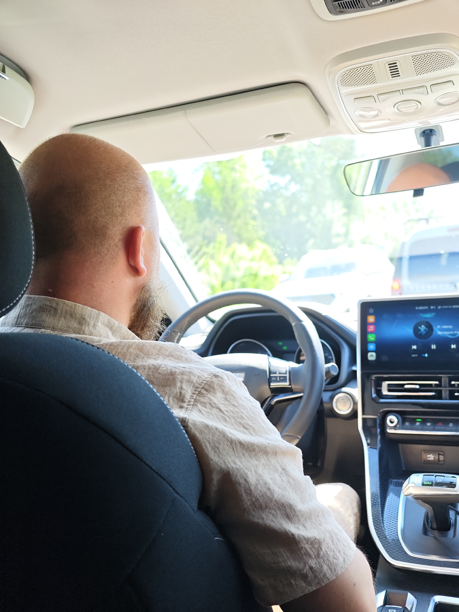 A person is seated in the driver's seat of a vehicle, facing forward. The individual has a bald head, a beard, and is wearing a light-colored, button-down shirt. The vehicle appears to be modern, with a large touchscreen display in the center console. The gear shift is visible, indicating the vehicle is likely an automatic transmission. Through the windshield, other vehicles and greenery suggest the scene is taking place outdoors. There are no distinctive characteristics in the surrounding environment to specifically suggest it is in Chaikovsky, Russia.