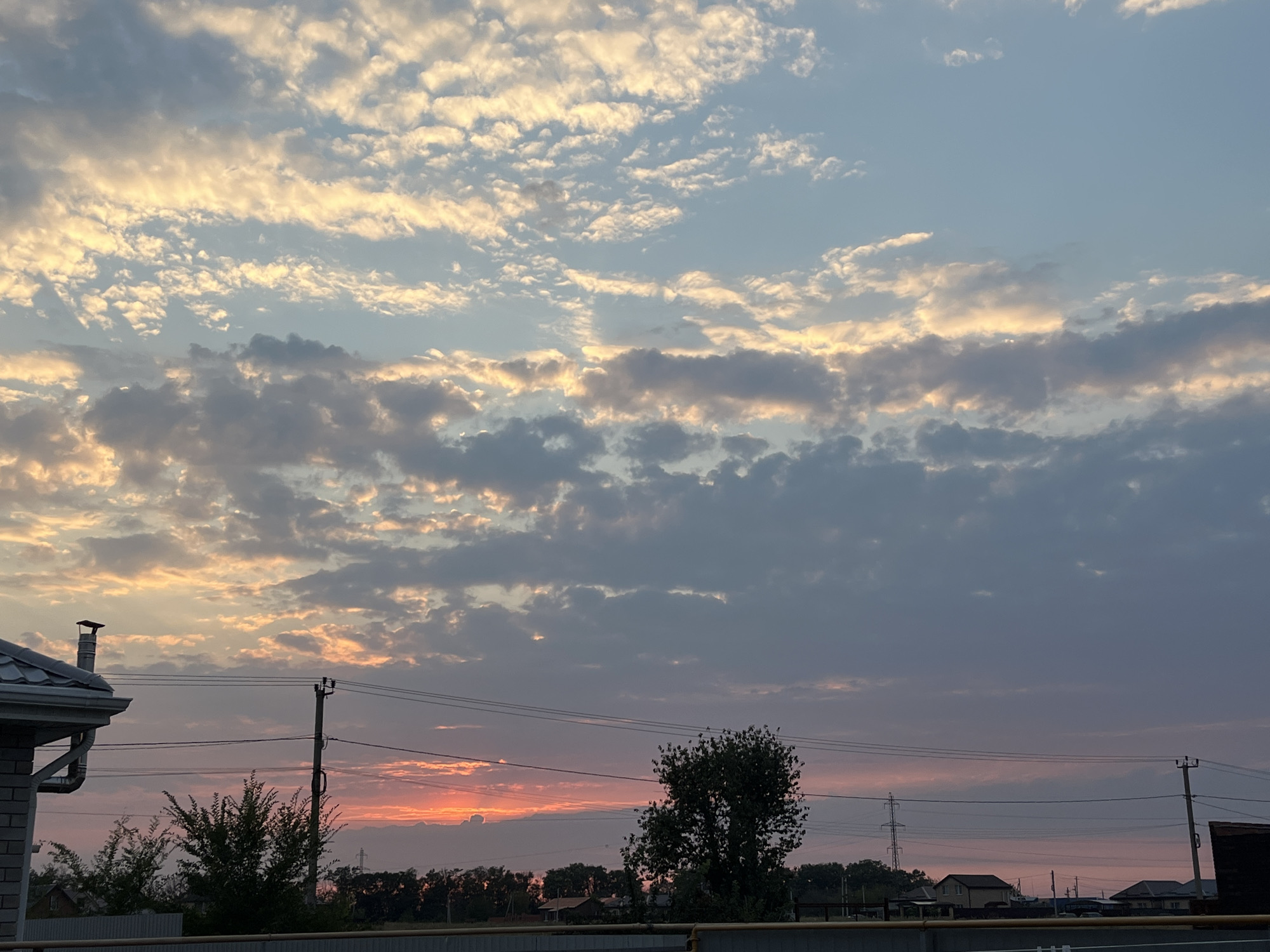 The image captures a sunset over a rural or suburban area. The sky is filled with dramatic clouds, tinged with orange, pink, and yellow hues from the setting sun, contrasting with the deeper blues and grays of the upper atmosphere. In the foreground, a section of a house with a gray roof and a chimney is visible on the left. Power lines and poles crisscross the middle ground, interspersed with trees and vegetation. In the distance, several houses and structures can be seen amongst more trees, suggesting a residential area. The overall impression is one of a peaceful evening scene in a location that appears to be outside of a major urban center, possibly within the Taganrog City District, Russia.