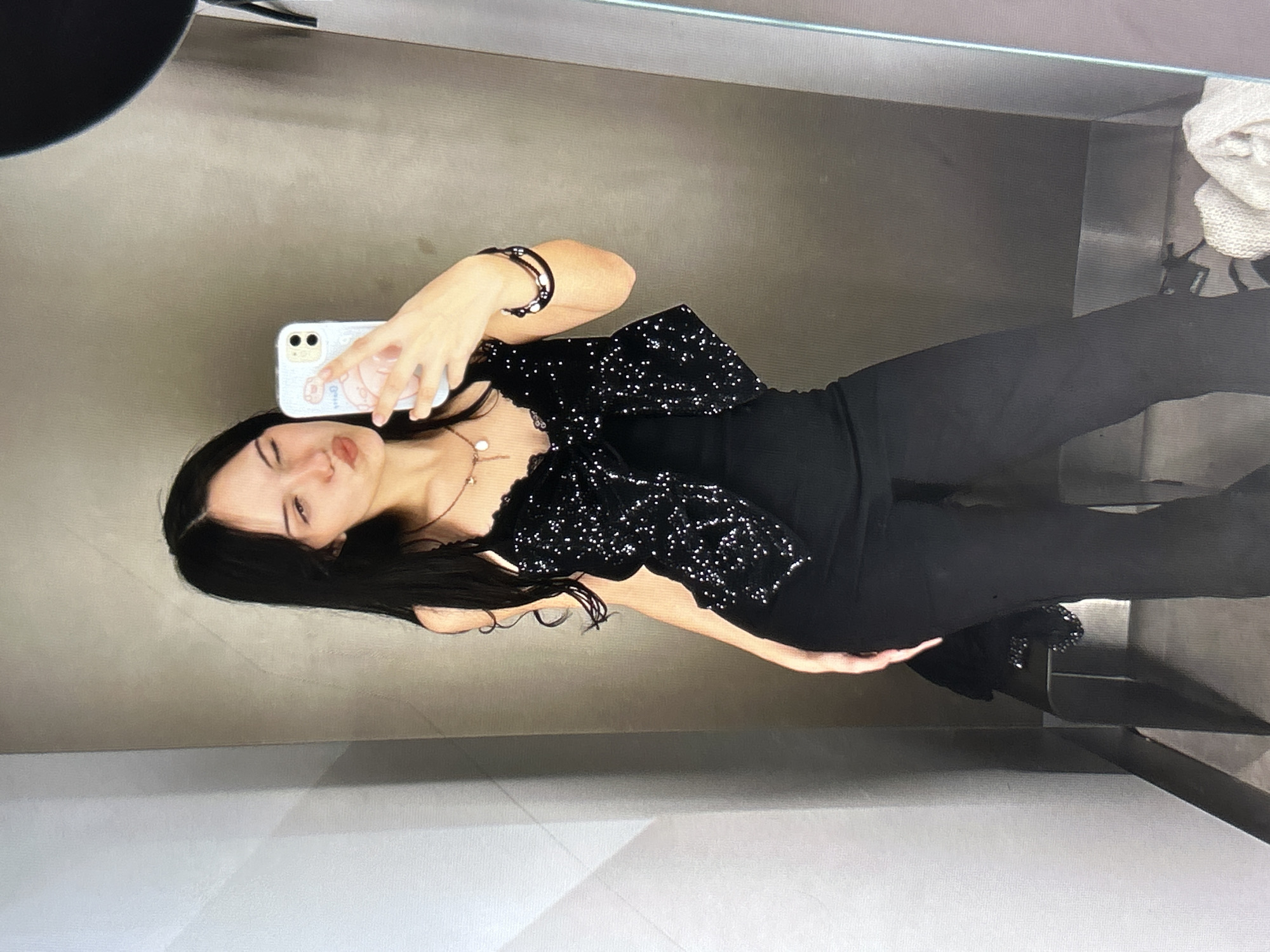 A young woman with long, dark hair is taking a mirror selfie in what appears to be a fitting room or a dimly lit space with textured walls. She is wearing a black, sequined top over a black tank top, and black flared pants. Her left eye is partially closed, and her lips are pursed. She is holding a light-colored phone with a cartoon case in her right hand, and her left hand is partially visible. A necklace with a pendant is visible around her neck, and she is wearing two bracelets on her right wrist. The background is a mottled gray wall, and a large black object, possibly a part of the mirror frame, is visible in the top right corner. The lighting is dim and casts subtle shadows. This scene does not have any discernible connection to Cupra Marittima, Italy.