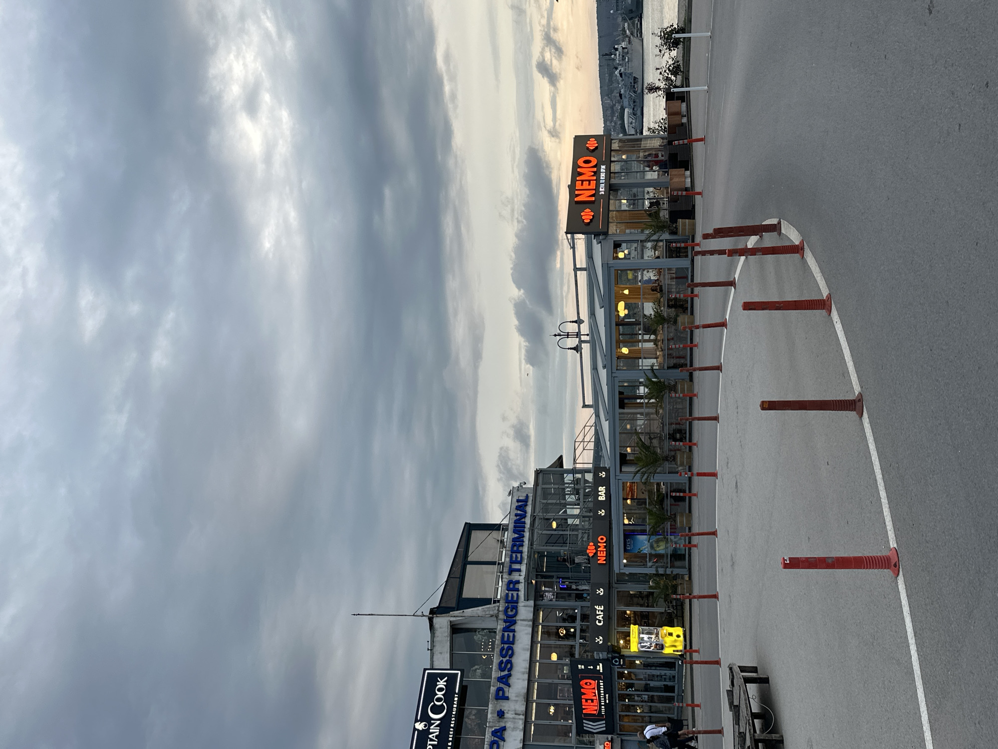 This media file depicts a passenger terminal building situated in Varna, Bulgaria, likely near a harbor or waterfront. The image is taken from the perspective of an adjacent asphalt area, possibly a road or parking lot. The sky is heavily overcast with dense clouds, suggesting a cloudy day or late afternoon/early evening light filtering through.
The main subject is a modern, multi-story building identified as a "PA PASSENGER TERMINAL." Several businesses operate within or next to it, prominently featuring bright orange neon signs. Visible text on the building includes "CAPTAIN COOK FISH & BEEF RESTAURANT" and multiple instances of "NEMO," associated with a "FISH RESTAURANT," "CAFÉ," and "BAR." One Nemo sign also mentions "300 M. FROM CITY CENTER" and "OPEN ALL DAY." The word "EFES" (a beer brand) is also visible.
In the foreground, a curved white line marks a section of the asphalt, bordered by a series of red and orange traffic cones or bollards. To the left, a wooden bench is partially visible, with what appears to be one or two individuals seated. In the background, beyond the building and across the water, a distant coastline with buildings can be seen, indicating a body of water such as the Black Sea or a bay. The overall atmosphere is functional and commercial, typical of a port area.