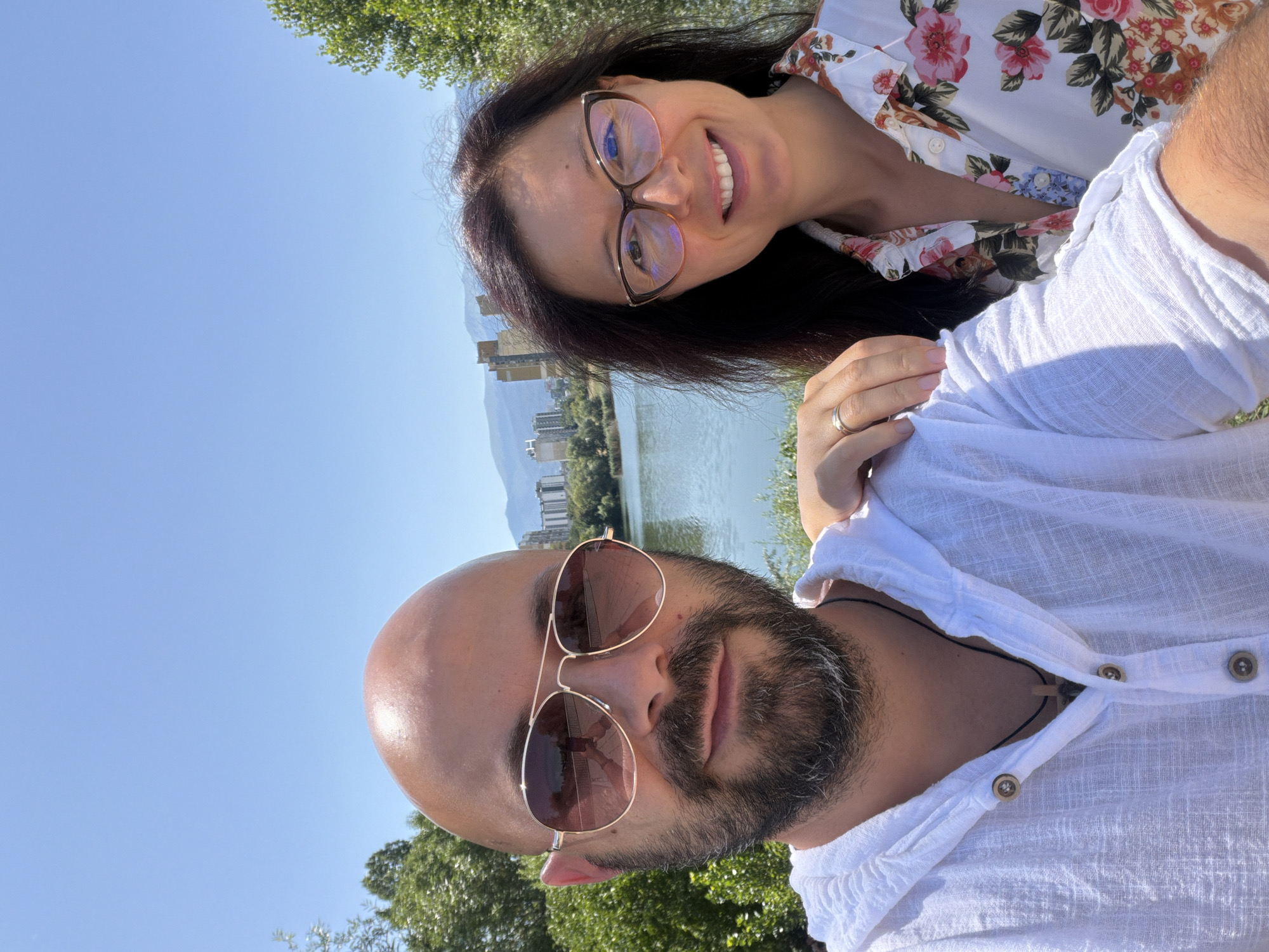 The image is a selfie taken outdoors on a sunny day, likely near a body of water with buildings and mountains in the background. A man with a bald head, beard, and sunglasses is in the foreground, wearing a white hooded shirt. Next to him is a woman with long dark hair, wearing glasses and a floral print shirt. The man's hand is resting on the woman's shoulder, and the woman has a ring on her left hand. The environment appears to be a park or recreational area with trees and a lake. The buildings in the background suggest an urban setting. The overall scene depicts a couple enjoying a day out.
Based on the presence of the lake and buildings, this scene is likely located in the area of the **Lake Ariana** or **Lake Pancharevo** in Sofia, Bulgaria. These locations are known for their natural beauty and recreational facilities within the city. The man's attire and the general ambiance suggest a casual outing.