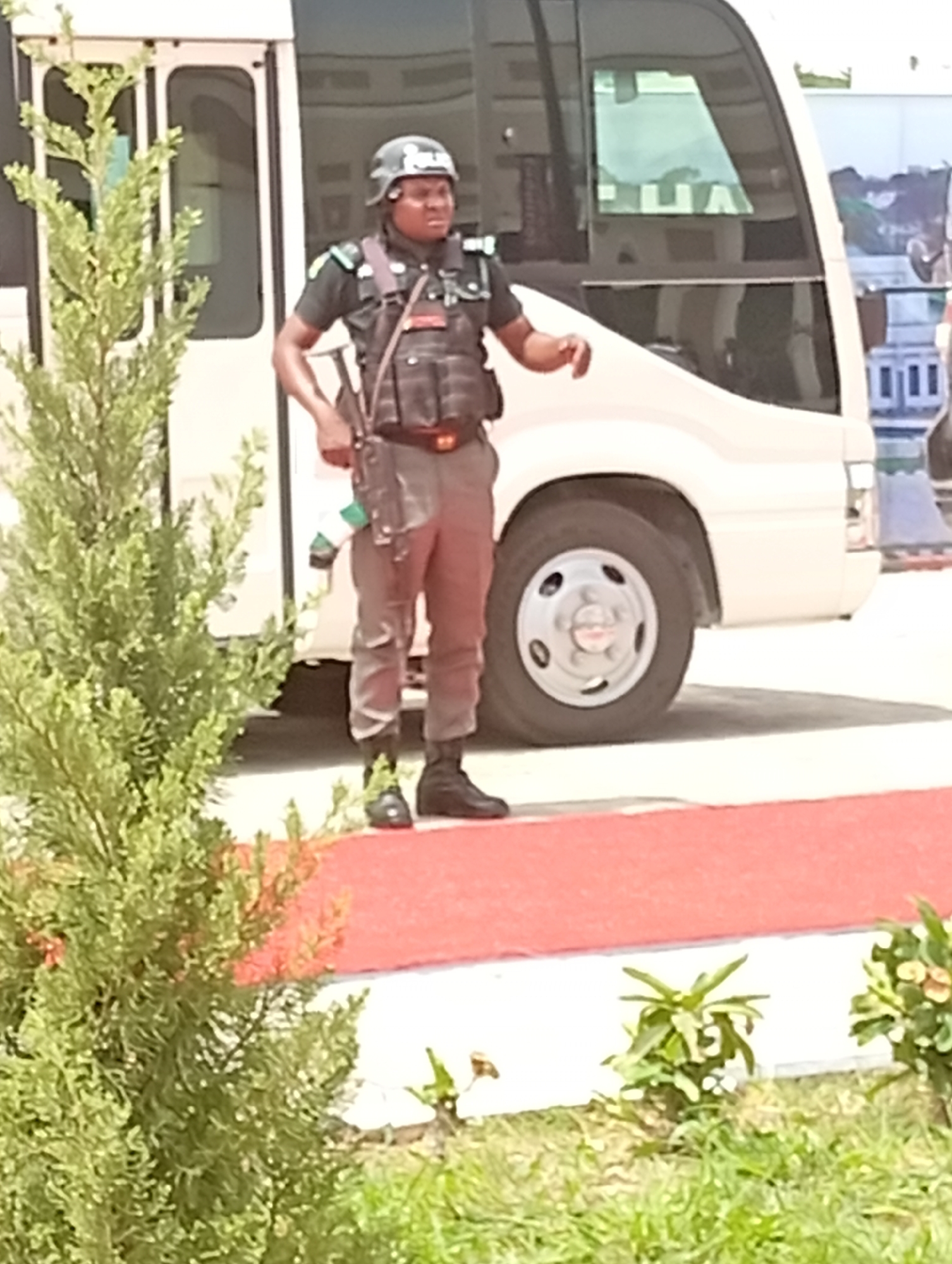 A security officer stands prominently in the foreground, dressed in a dark uniform consisting of a short-sleeved top, tactical vest, pants, and black boots. They wear a dark helmet and have straps across their chest and shoulders, with what appears to be a small white and green emblem or flag hanging from a holster on their right hip. The officer is looking towards the right side of the frame with a focused expression, their right arm slightly raised.
Behind the officer and to their right is a white mini-bus or coaster bus, partially visible. The front section of the bus, including a headlight, part of the windshield, and a wheel, can be seen. Faint reflections on the bus window show what might be inverted text, possibly "HA".
The ground in front of the officer is covered by a red surface, resembling a carpet or painted pathway, which transitions to a lighter, possibly paved, area. In the foreground and to the left, a tall, dense green shrub or tree partially obstructs the view. Smaller green plants and grass are visible at the very bottom of the frame.
The scene takes place outdoors during the daytime, under bright lighting, suggesting clear weather. The officer appears to be on duty, possibly guarding the area or observing. Given the context of Unguwan Sanusi, Nigeria, the uniform and the green/white emblem are consistent with Nigerian security forces.