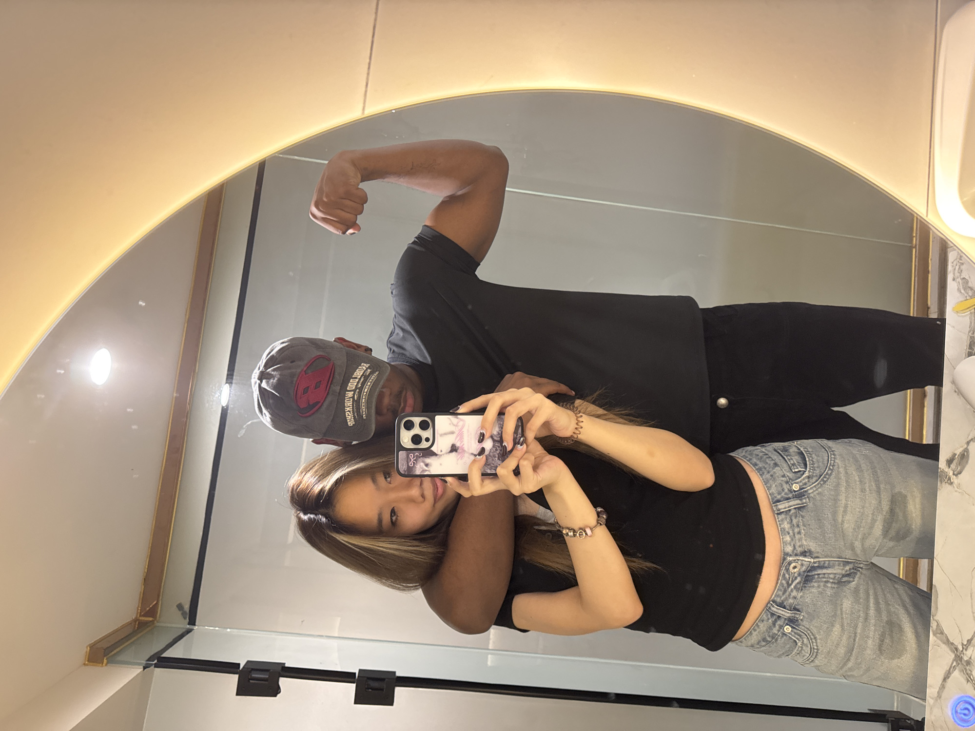 A couple poses for a selfie in a bathroom mirror. The man, wearing a black shirt and a gray baseball cap with a red logo, flexes his arm. The woman, with long brown hair, holds up a phone with a patterned case to take the picture. She is wearing a black top and light blue jeans. The bathroom has a large, round mirror with a lighted border. The location is implied to be in the city of Yuzhong District, China.