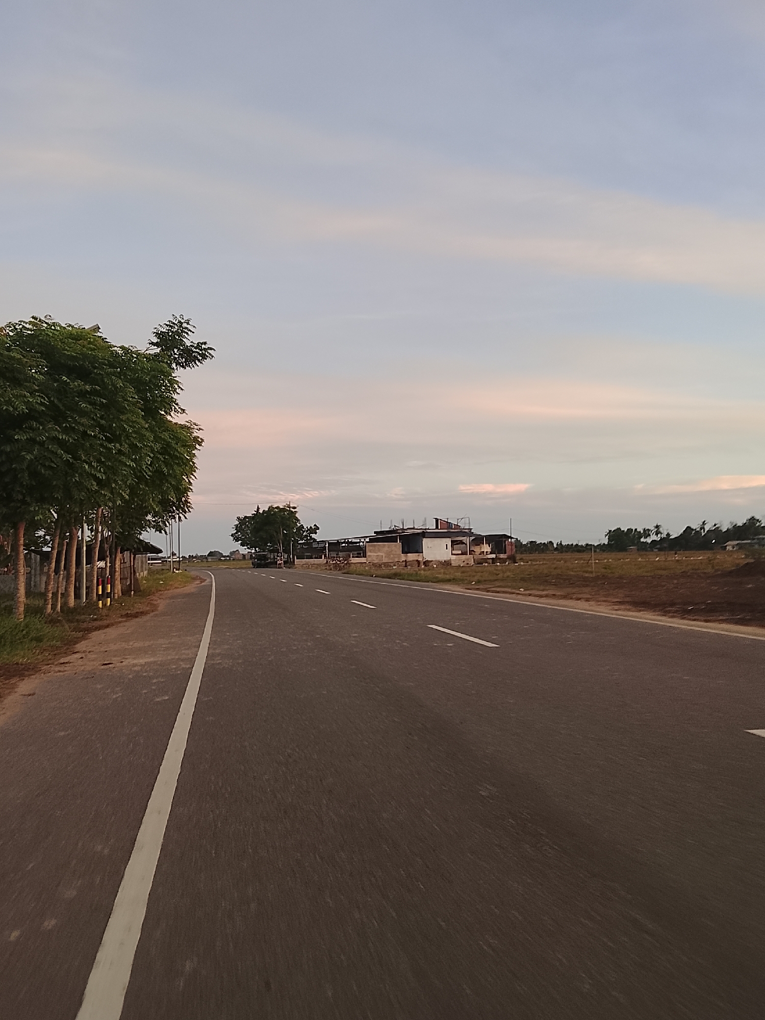 The image shows a paved road with a white line marking its left edge, curving to the right. The road is bordered on the left by a row of tall, leafy green trees and a low fence. In the distance, across a field, are several buildings under construction, with exposed framing and concrete work. The sky above is a pale blue with soft, pinkish clouds, indicating either sunrise or sunset. The general environment appears rural or semi-rural, with open fields and scattered structures. The location is the city of Dayah Kruet, Indonesia.