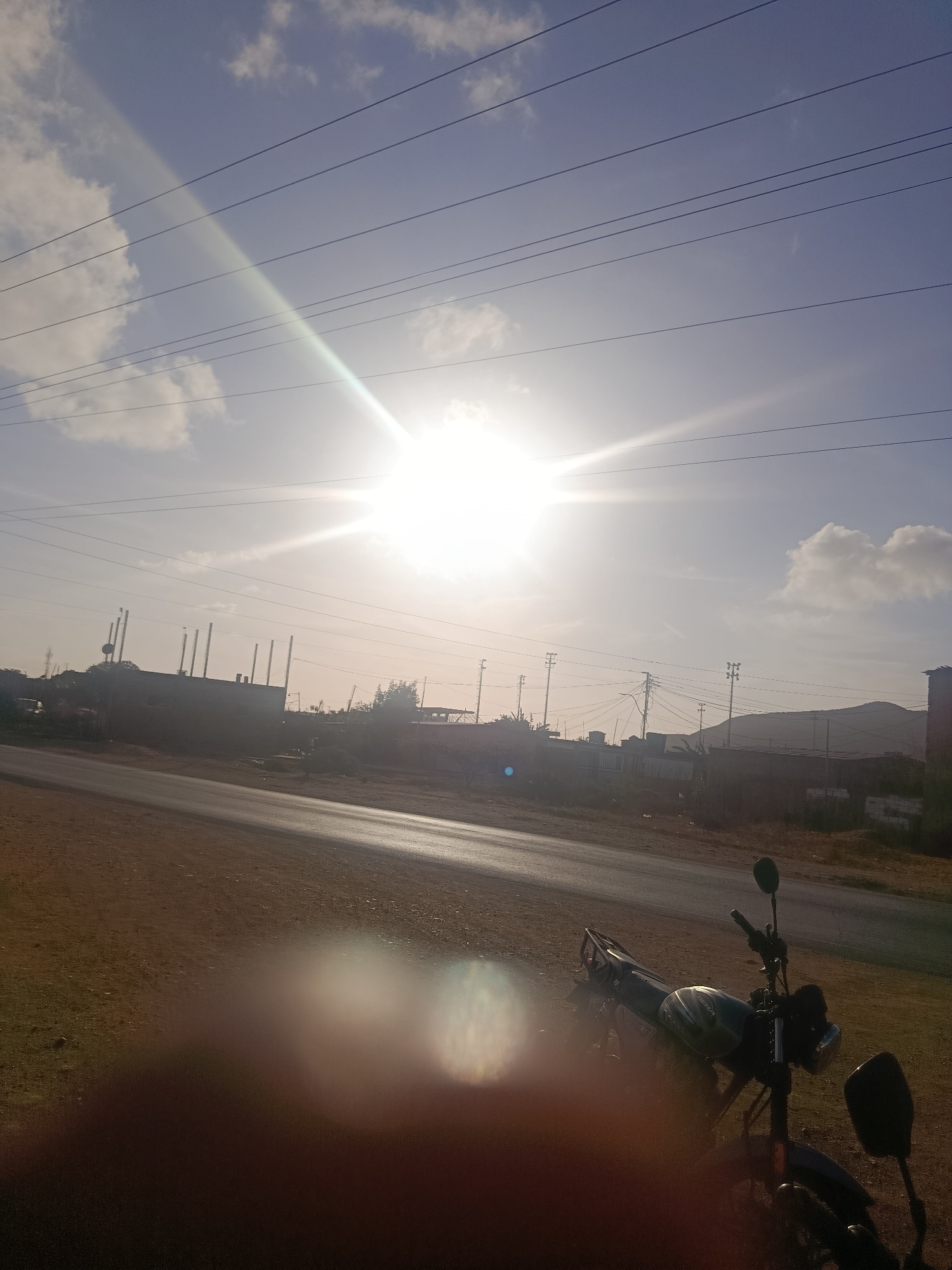 The photo, taken in the city of Juan Griego, Venezuela, shows a bright, sunlit scene with a motorcycle in the foreground. A road runs horizontally across the middle ground, with a blurred shape possibly a hand in the lower portion of the photo. Beyond the road, buildings and structures are visible against a backdrop of hills. Power lines stretch across the upper portion of the sky, punctuated by scattered clouds. The overall lighting is harsh due to the direct sunlight.