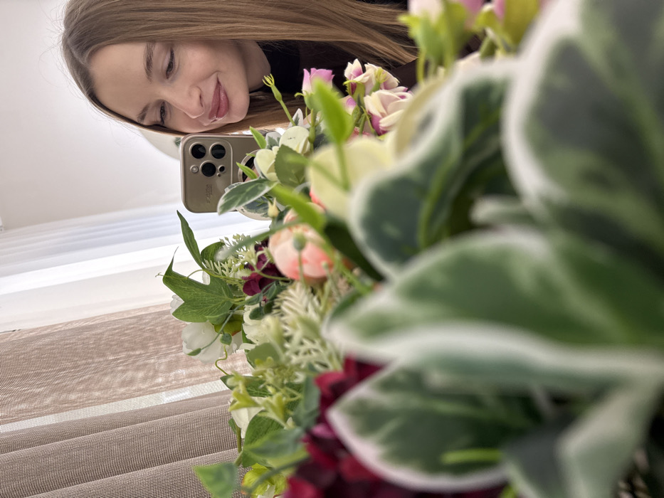 The image presents an indoor scene featuring a woman partially obscured by a foreground floral arrangement. On the right, a woman with long, straight light brown hair, wearing a dark high-necked garment, looks towards the left with a subtle smile. She holds a light gold/tan smartphone, equipped with a prominent multi-lens camera system, reflecting her image, indicating a selfie or mirror photograph. The foreground and central-left portions of the frame are occupied by a large, out-of-focus bouquet or arrangement of flowers and foliage, including white peony-like blossoms, small pink flowers, deep red petals, and various green leaves, some variegated with white edges. In the background, behind the woman and the flowers, sheer brown and white curtains are visible, indicating a window and a source of natural, diffused light. The setting is generic and lacks specific architectural or environmental markers to identify a precise location within Budva, Montenegro.