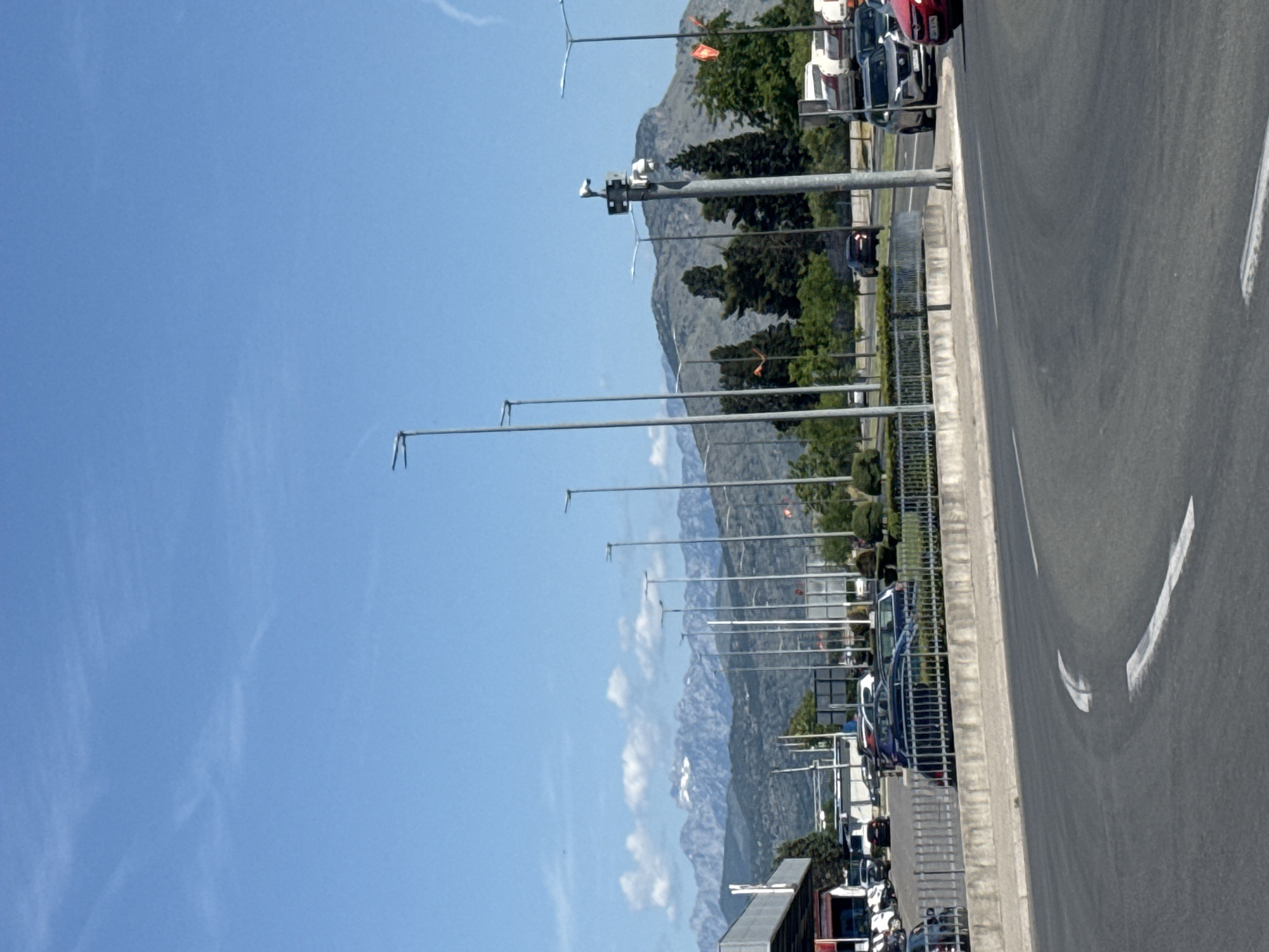 The image depicts an outdoor scene featuring a multi-lane asphalt road curving to the left in the foreground, marked with two white arrow-shaped road markings. To the immediate right of the road is a low concrete barrier topped with a grey metal fence, behind which a parking area is visible with multiple parked vehicles including dark sedans and light-colored SUVs. Further back on the left, an architectural structure with a wide, light-colored roof overhang is partially visible, and several white scooters or motorcycles are parked nearby.
Numerous tall, grey metal streetlights, each with an L-shaped arm holding a light fixture, line the road. In the mid-ground, on the right side of the road, a robust grey utility pole is equipped with at least two surveillance cameras, one facing upwards and one downwards, along with other technical components. Behind the fence and streetlights, various green trees are present, including dense, dark-foliaged conifers. Several small, red or orange flags are attached to poles in the mid-ground, partially obscured by foliage.
In the distance, a prominent range of rugged mountains with visible patches of white, possibly snow or light-colored rock, dominates the left-to-center horizon. To the right of this range, a large, less rugged mountain with sparse vegetation is visible. The sky is clear blue with a few scattered, wispy white clouds, primarily above the distant mountains on the left. A faint white streak, possibly a contrail, is visible in the upper right quadrant of the sky. On the road, a dark-colored SUV is visible in the distance, and closer to the foreground on the right, a silver SUV and a partially visible red car are present. Several white semi-trailer trucks are parked in a lot further back on the right. No people are discernible. The scene is brightly lit, suggesting daylight. The location is the city of Unknown, Unknown.