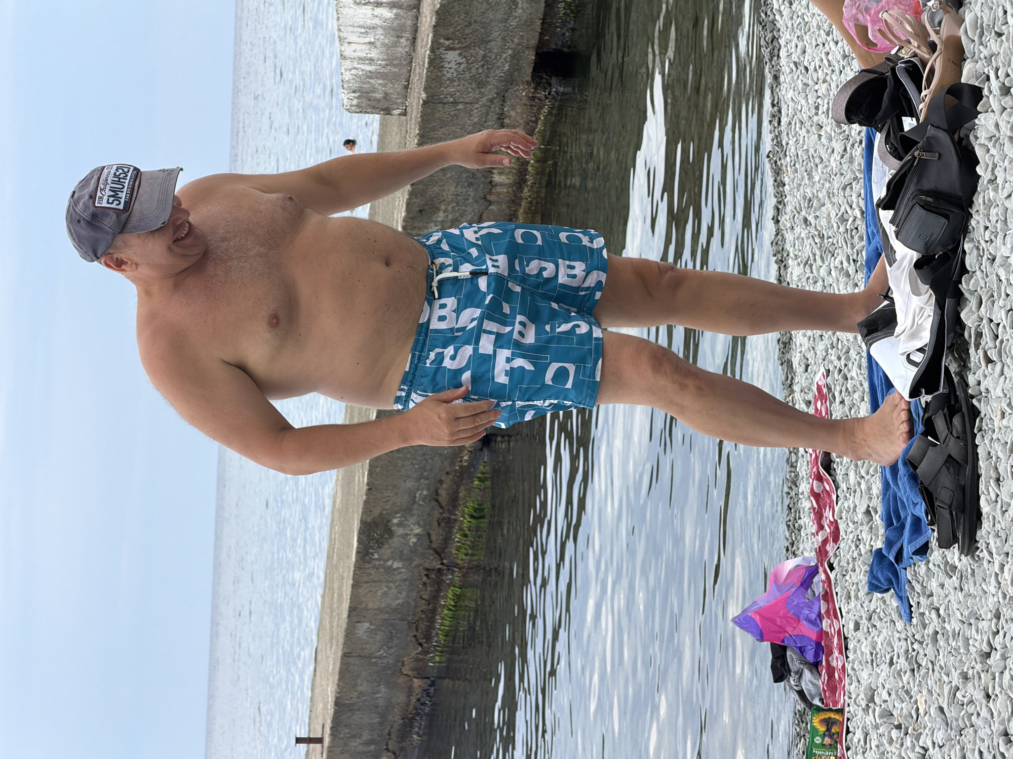 An overweight, bald, white man wearing a grey baseball cap and teal swim trunks stands on a pebble beach. He is looking down and laughing, with his right arm bent and his hand on his hip. The man is standing in front of a concrete pier with a calm sea beyond. There are several items of clothing and a bag on the beach near the man's feet. The location is the city of Partenit, Ukraine.