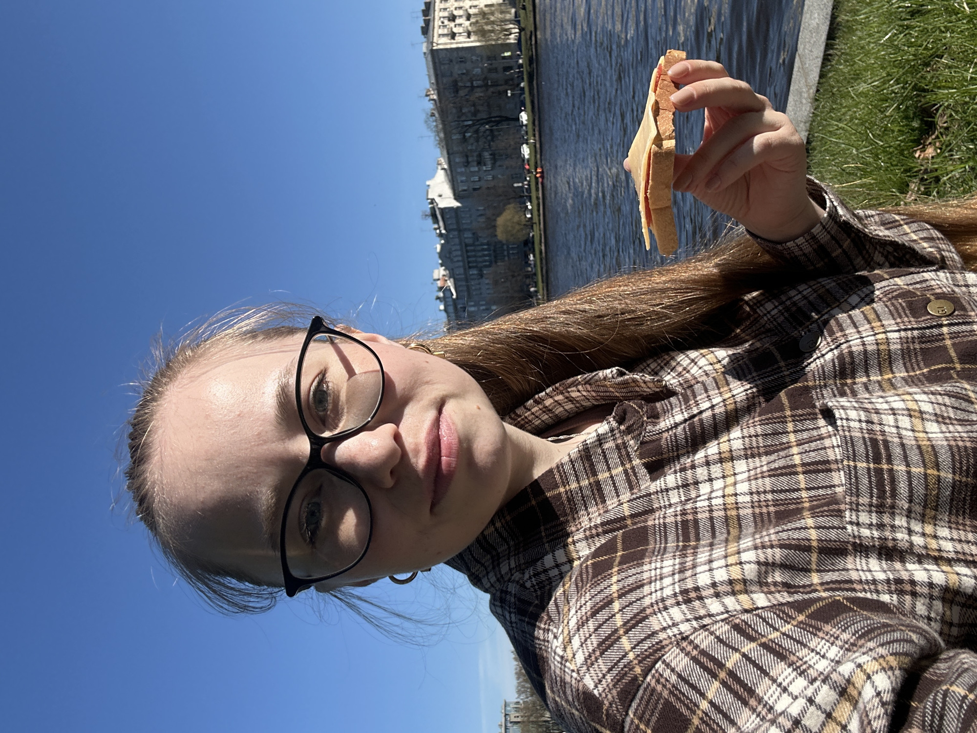 A young woman is holding a sandwich, possibly in Saint Petersburg, Russia. She is wearing glasses and a plaid shirt. Behind her is a river and city buildings under a blue sky. The picture appears to be a selfie.
