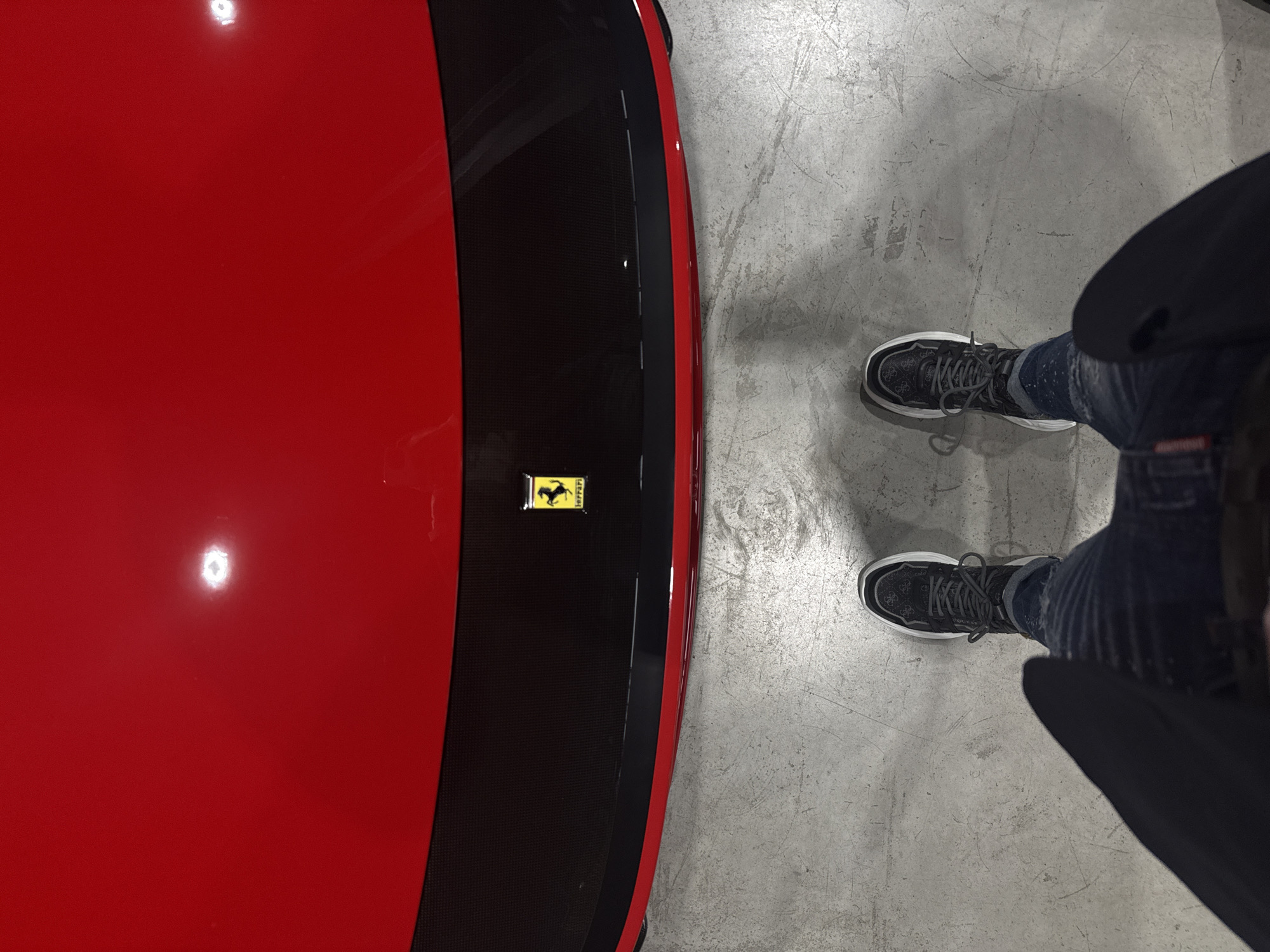 The image, captured from an overhead perspective, features a person's lower body standing beside a vibrant red car in what appears to be an indoor setting, likely a garage or showroom in Plovdiv, Bulgaria. The left side of the frame is dominated by a glossy, highly reflective red surface of the car, possibly the hood or trunk. Running down the center of this red body is a strip of black, possibly carbon fiber, on which a small, iconic yellow rectangular Ferrari emblem is affixed, displaying the black prancing horse and the text "FERRARI" above it.
To the right, a person is visible from the waist down, wearing dark blue jeans that show signs of distress or white paint splatters. They are sporting dark grey or black sneakers with prominent white soles. An intricate pattern is visible on the side of the sneakers, where the brand name "GUESS" is clearly discernible. The ground beneath is a light grey concrete floor, appearing textured and scuffed, indicative of an industrial or vehicle display environment. Artificial lighting is suggested by the bright reflections on the car's polished surface. The scene conveys a casual interaction with a luxury automobile.