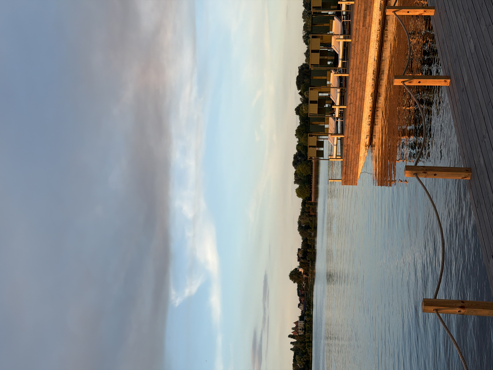 This media file depicts a serene waterside scene, likely at dusk, given the warm, golden hues reflecting on the water and the soft lighting. The perspective is from a wooden deck, which occupies the right side of the frame, extending towards the horizon. Along the edge of the deck, several lounge chairs with muted green and yellow cushions are arranged, suggesting a recreational or hospitality setting.
Beyond the deck and chairs, a calm body of water stretches out, with gentle ripples disturbing its surface and reflecting the sky and the distant shoreline. The shoreline is lined with lush green trees and dotted with houses, indicating a residential area or a park bordering the water.
The sky dominates the left side of the image, transitioning from a soft, pale blue near the horizon to a more muted, overcast grey towards the top. Wispy clouds are present, some catching the subtle sunlight.
The overall atmosphere is peaceful and tranquil, evoking a sense of relaxation and quietude. The wooden elements of the deck and the rope detailing with wooden posts add a rustic touch to the scene. The time of day appears to be late afternoon or early evening, with the sun casting long shadows and a warm glow. No people are visible in the frame, and no text is present. The location context provided places this scene in Kyiv, Ukraine, suggesting it might be a view of the Dnieper River or a similar waterway within the city.