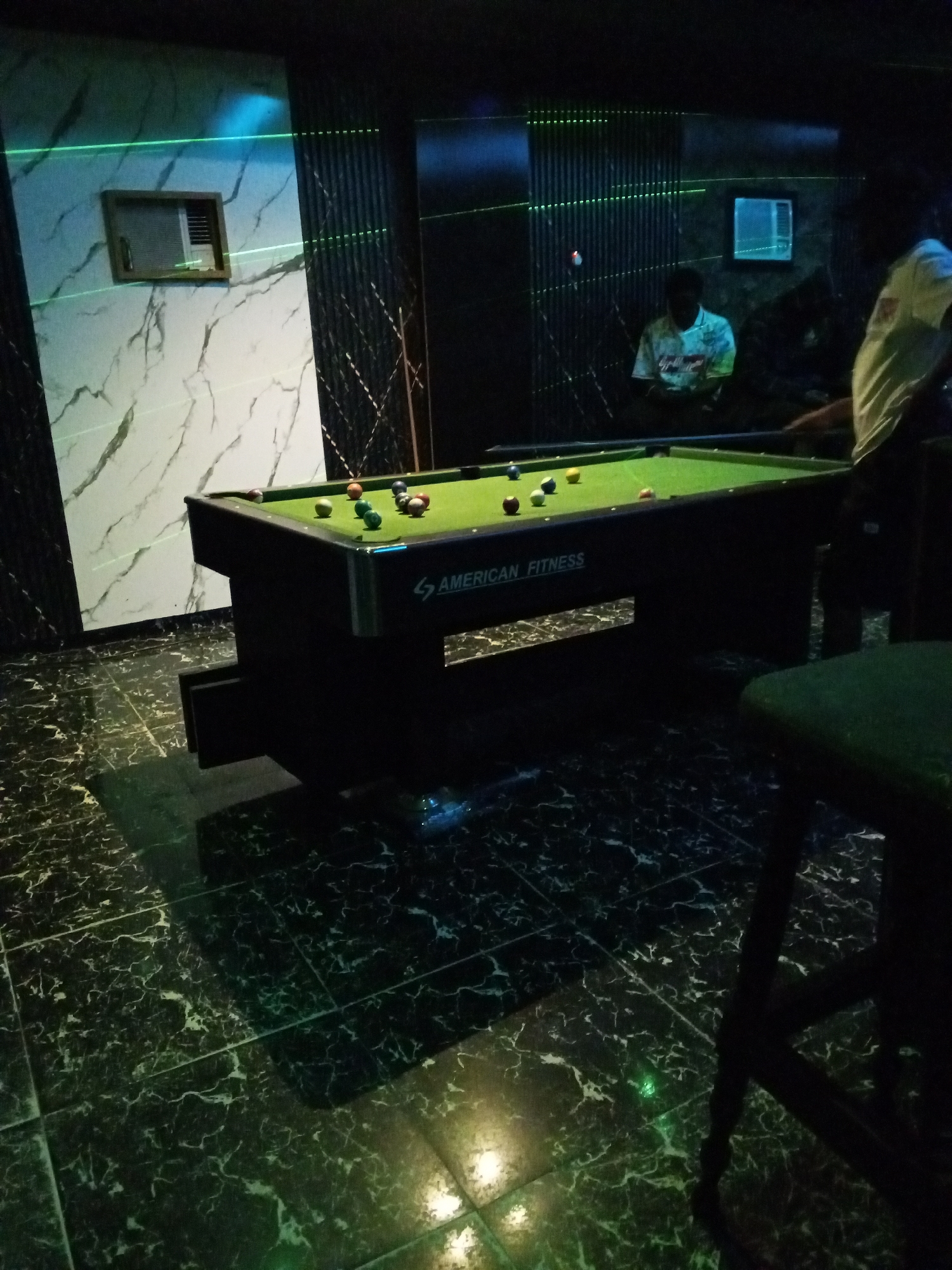 The image depicts an indoor billiard hall, likely in Lokoja, Nigeria, given the provided context. A pool table with multiple balls is the central focus, set against a backdrop of dark walls with striking green laser lines and a marble-patterned wall. Two individuals are visible: one in the background, wearing a patterned shirt and looking towards the table, and another on the right, partially visible and appearing to be playing. The floor is tiled with a dark marble pattern that reflects the ambient lighting. The overall atmosphere suggests a recreational setting.