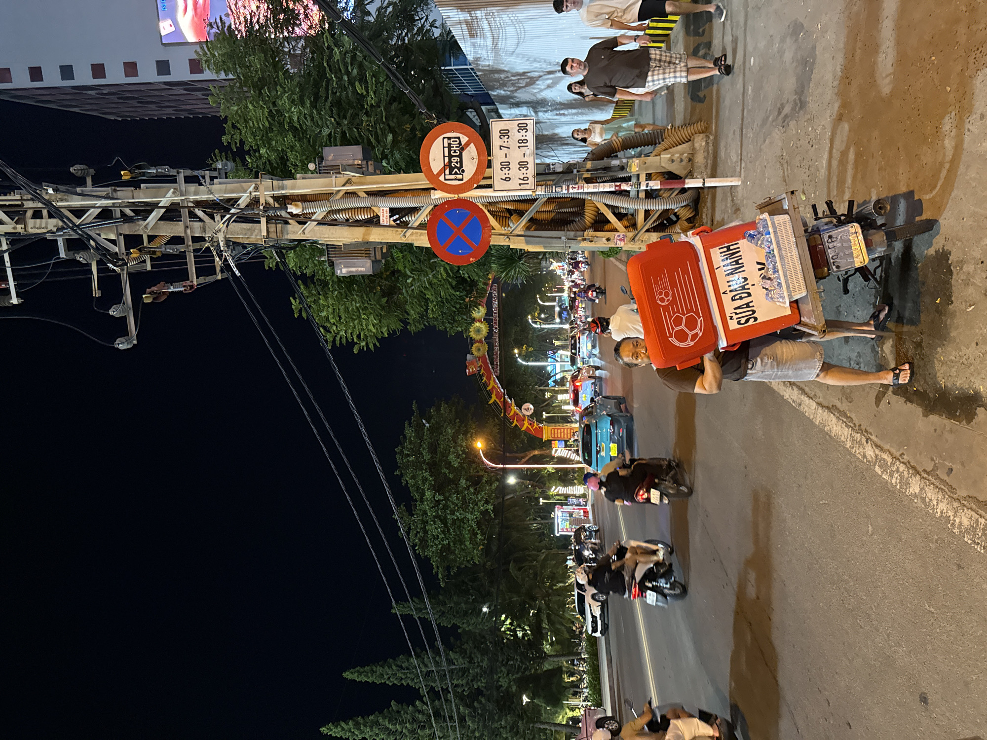At night on a street in Nha Trang, Vietnam, a man stands beside a motorcycle carrying a red cooler box filled with bottled drinks. The motorcycle has a license plate that reads "79F5 01763". The cooler box has Vietnamese text that translates to "Soybean Milk". In the background, traffic including cars and motorcycles moves along the street. Pedestrians walk on the sidewalk. A traffic pole with various signs and electrical wires is visible on the right side of the image. One sign indicates parking restrictions between 6:30-7:30 and 16:30-18:30. Trees and decorative arches are also present.