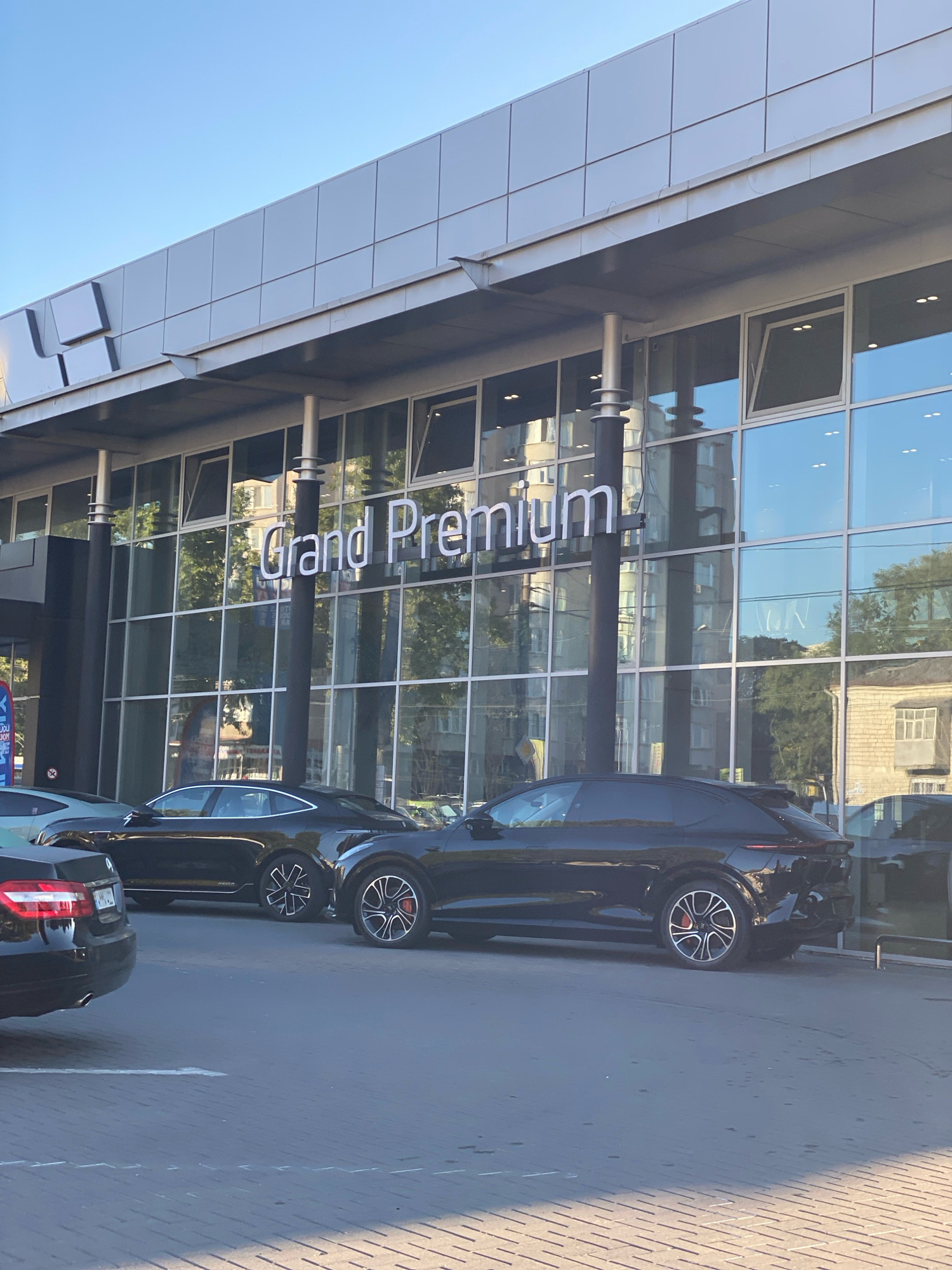 The image shows the exterior of a car dealership named "Grand Premium" in Chișinău, Moldova. The dealership has a modern facade with large glass windows and silver paneling. Several cars are parked in front of the building, including a black SUV, a black sedan, and a portion of a silver car. The sky is clear and blue. The scene appears to be during daylight.