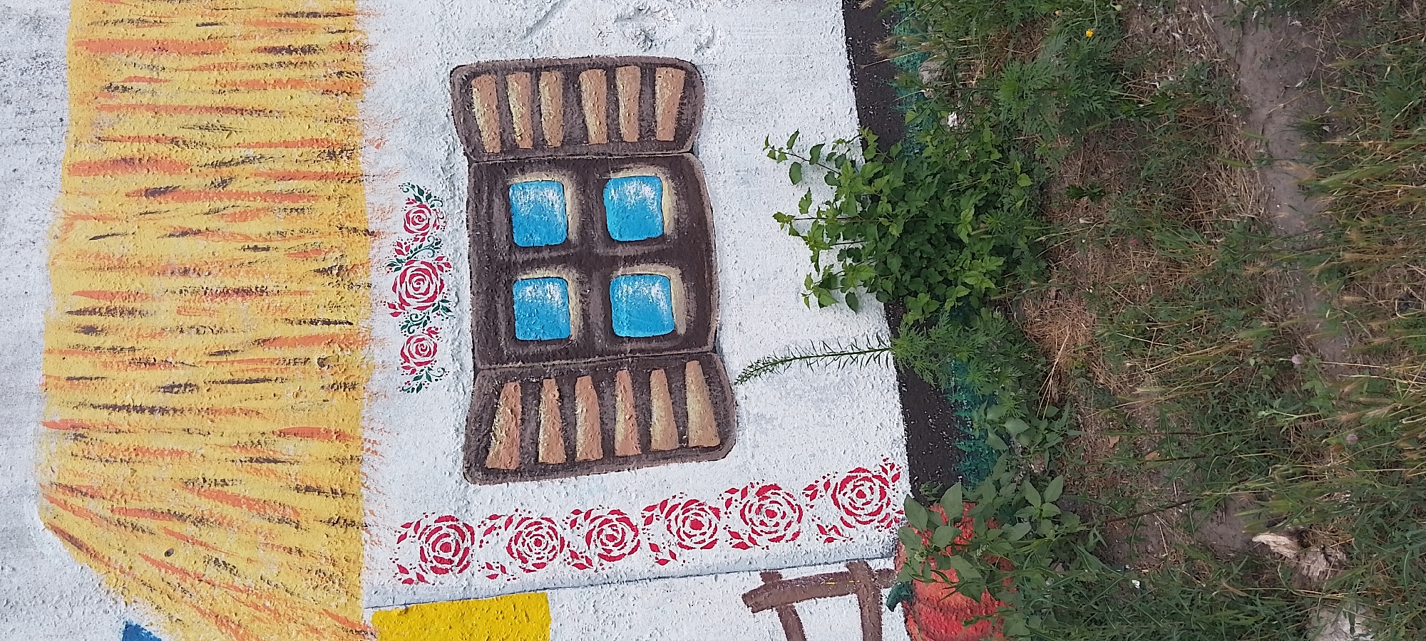 The image displays a mural painted on a concrete wall. The mural depicts a stylized representation of a traditional house with a thatched roof. The roof is painted in shades of yellow and orange, with vertical brushstrokes suggesting straw. Below the roofline, the wall is white and features a decorative border of red roses on the left side. A brown window with four blue panes is centered on the white wall, with wooden shutters painted on either side. Above the window, there is a smaller floral motif of red roses. To the left of the window, a portion of a red pumpkin is visible, along with a wooden fence post. In front of the mural, a patch of overgrown green vegetation and dry grass is present. The vegetation includes various plants and weeds. The ground appears to be uneven, with patches of dirt and dry soil. The overall scene suggests an outdoor setting, possibly a courtyard or the side of a building in a rural or suburban area. The presence of the mural indicates an artistic or decorative element applied to the wall. The location is not explicitly stated in the image, but the prompt specifies that the image is from Cherkasy, Ukraine.