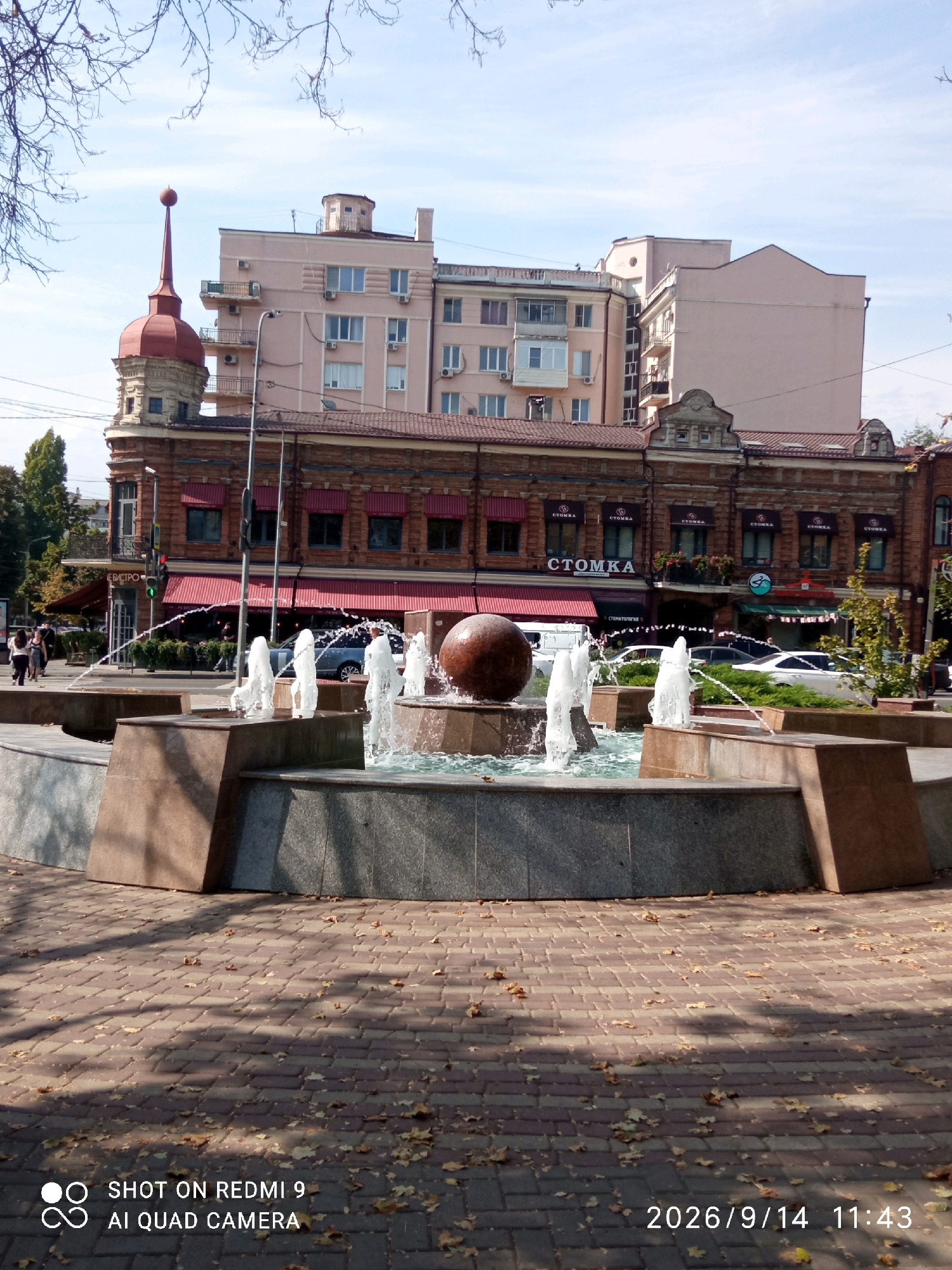 A fountain with spouting water is in the foreground of the image. Behind the fountain, a brick building with red awnings is visible. Several people are walking in front of the building. The building has several windows and a sign that reads "СТОМКА". In the background, there are other buildings, including a tall, light-colored building with a red turret. The sky is blue with some clouds. The ground in front of the fountain is paved with bricks and fallen leaves. The image is dated 2026/9/14 11:43.