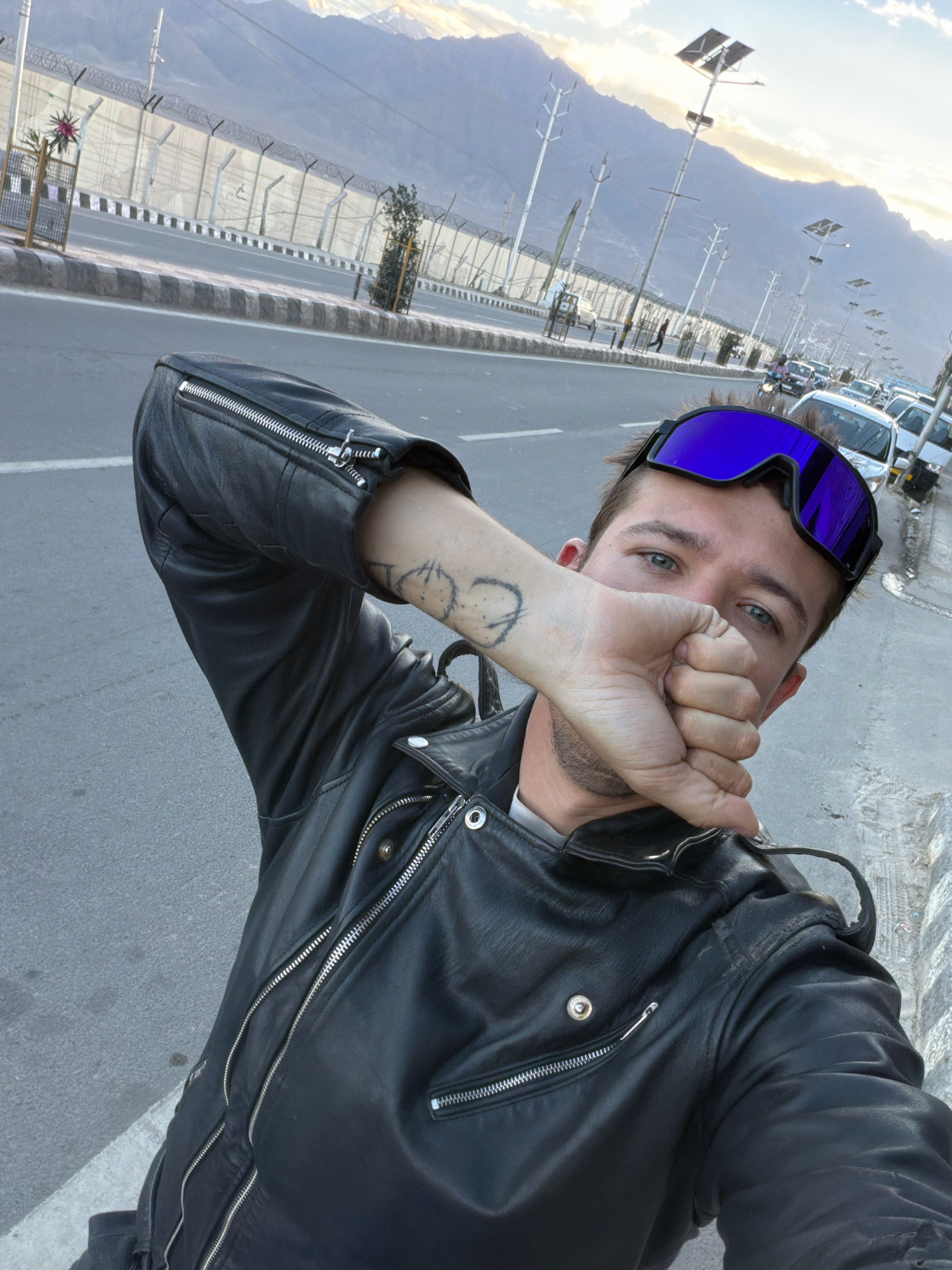 A man wearing a black leather jacket and blue ski goggles is taking a selfie on a street in Leh, India. The man's right hand is covering his mouth, and he has a tattoo on his left arm. In the background, there is a road with cars and motorcycles, a fence, and mountains. The sky is cloudy with the sun setting.