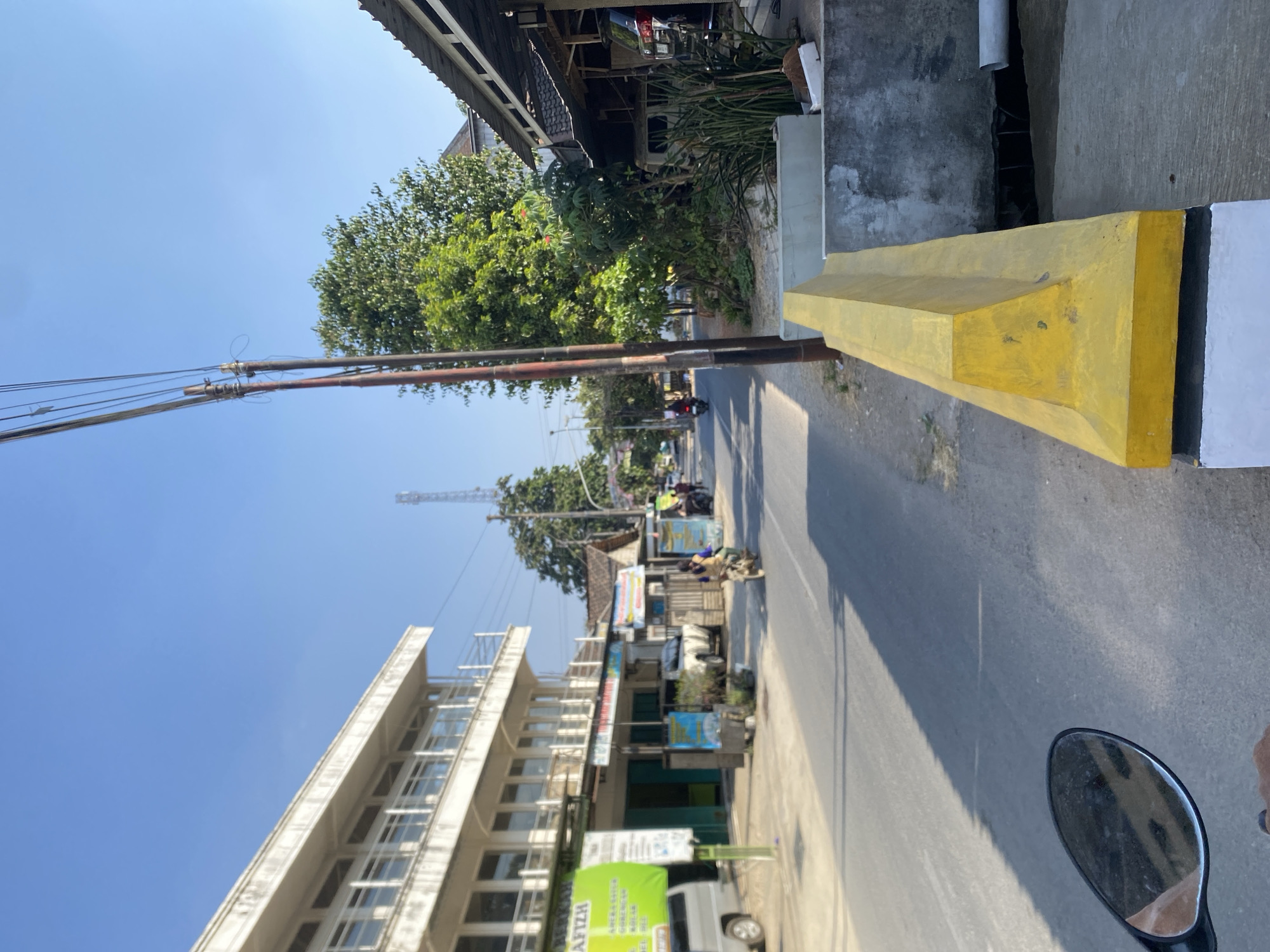 The image captures a street scene on a clear day in an urban environment. In the foreground, a yellow concrete barrier is prominently featured, suggesting it might be a median or a roadside divider. To the left, a motorcycle's side mirror, reflecting a hand and the road ahead, indicates the viewpoint of someone on a motorcycle. The road surface is asphalt, with shadows cast from the surrounding structures and a utility pole.
On the left side of the street, a multi-story white building with multiple windows and balconies is visible. A green banner with text is attached to the lower part of this building. Several vehicles, including a silver van and other cars, are parked or moving on the street.
Further down the street, a variety of smaller buildings and shops line the right side, some with awnings providing shade. A person on a motorcycle is seen riding away from the camera. A tall communication tower is visible in the distance, against the clear blue sky. Lush green trees are scattered throughout the scene, adding to the natural elements within the urban landscape. The overall impression is one of a moderately busy street in a town or city.
