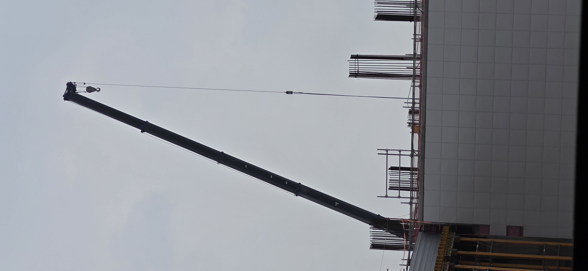 The image depicts a construction site in Pyeongtaek-si, South Korea. A large black crane arm extends diagonally across the frame, reaching upwards and to the right. A cable hangs from the crane, with a hook attached at the end. Below the crane, the upper section of a building under construction is visible. This section features a facade of light-colored, square tiles. Rebar structures are present on the roof of the building, along with scaffolding. The sky is overcast and gray. No people are visible in the image.