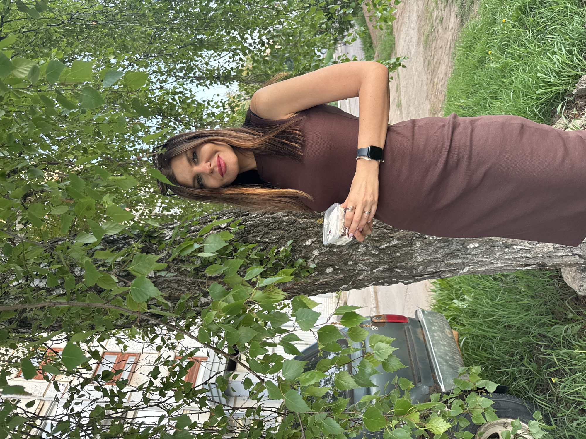 A woman in a brown dress is standing next to a birch tree. She is holding a small container. A green car is partially visible behind the tree. The location is Chaikovsky, Russia.