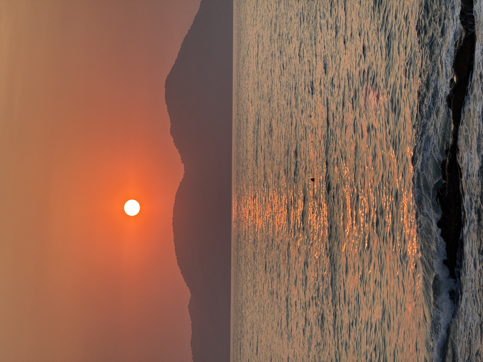 A serene sunset over the Aegean Sea, with the sun a bright white orb partially obscured by a hazy orange sky. In the distance, silhouetted mountains form a jagged horizon line. The calm water reflects the sun's golden glow, creating a shimmering path across its surface. Small waves gently lap against dark, rocky shores in the foreground. The scene is devoid of people and suggests a peaceful, natural environment. This is likely a view from Fethiye, Turkey, a coastal town known for its beautiful sunsets over the sea.