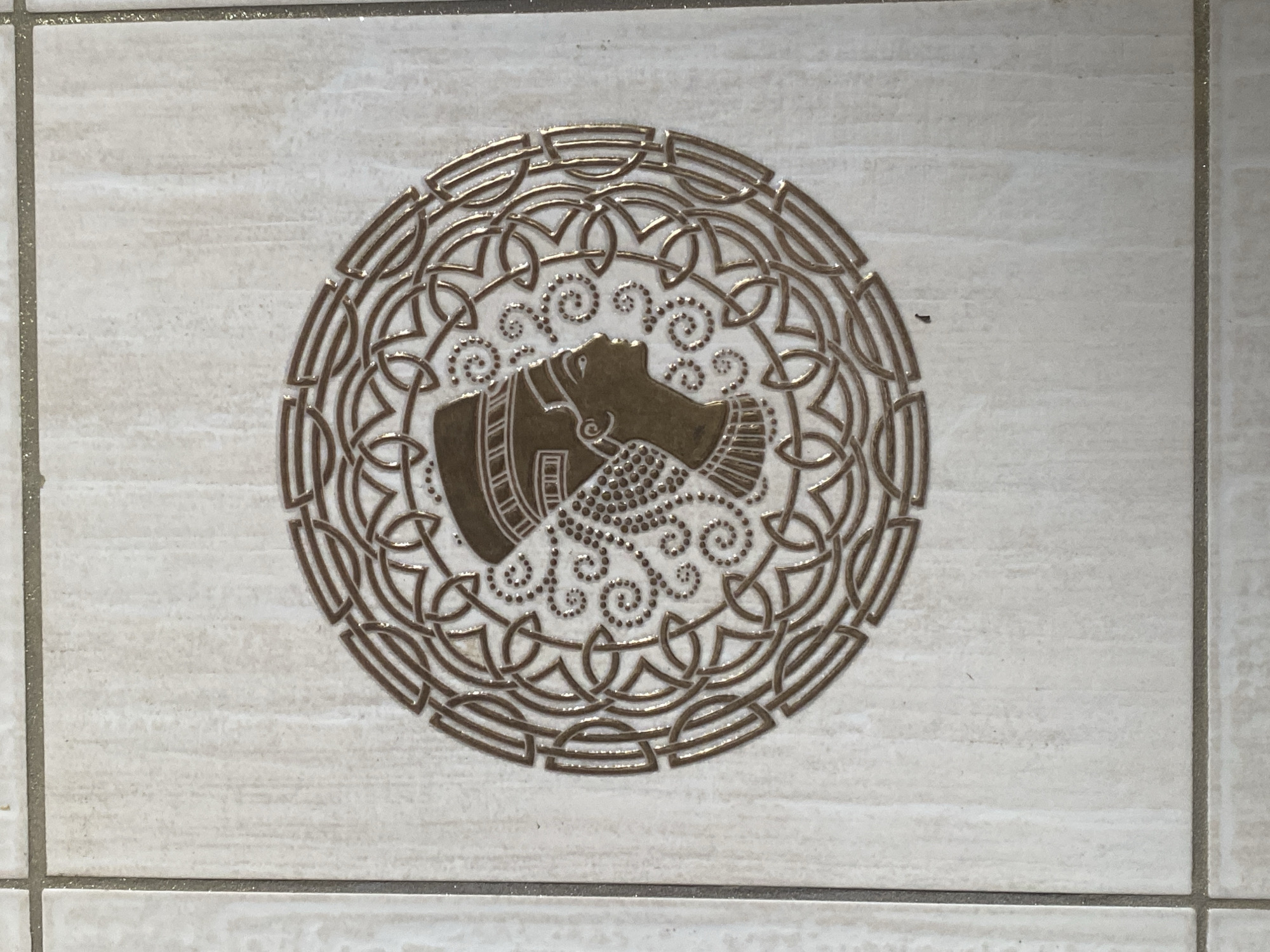 A close-up shot shows a square tile within a grid of similar tiles, likely forming part of a wall or floor. The tile features a metallic gold design on a light beige background with subtle horizontal striations. The design is primarily circular, consisting of an intricate pattern of interwoven lines that surround a central motif. The central motif is a stylized bust of a person in profile facing to the right. The person wears a decorative collar and headdress, with beaded strands or curls emanating from the headdress. The location is Dzerzhinsk, Russia.