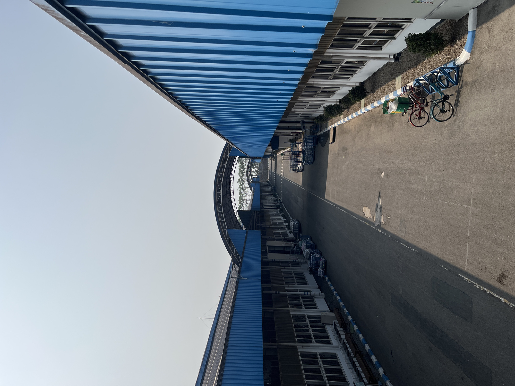 This image depicts an outdoor scene on a clear day, likely in an industrial or commercial area. The dominant structures are long, low-rise buildings with blue corrugated metal roofs and white walls. These buildings line a wide asphalt road or walkway, suggesting a factory, warehouse, or logistics complex. The architecture features numerous windows and what appear to be loading bays or entrances along the sides of the buildings.
In the foreground, a blue and white striped barrier runs along the edge of the road. Parked near this barrier are two bicycles, one red and one blue, next to a green trash bin. Further down the road, several multi-wheeled carts are visible, some laden with what appear to be packaged goods. The road itself shows some signs of wear, with patched areas and scattered debris.
In the background, the road curves and leads under a large, arched canopy structure, also with a blue roof. Beyond this, more buildings and trees are visible, stretching towards the horizon under a pale, hazy sky. The overall impression is one of an organized, functional outdoor space, possibly within a larger industrial park.
The image does not contain any explicit visual cues to pinpoint its exact location within Pelang, Indonesia, but the architectural style and climate suggest a tropical or subtropical setting consistent with Indonesia.