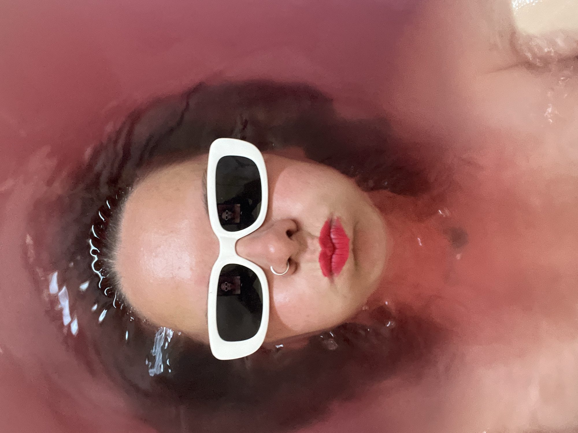 A person is submerged in a pink-tinted body of water, presumably a bathtub. The person is wearing sunglasses with thick white frames and has a nose ring. Their lips are painted red. The water is rippled and the person's hair appears wet and dark. The location is not discernible from the image and there is no information in the image to suggest it is taken in the city of Chaikovsky, Russia.